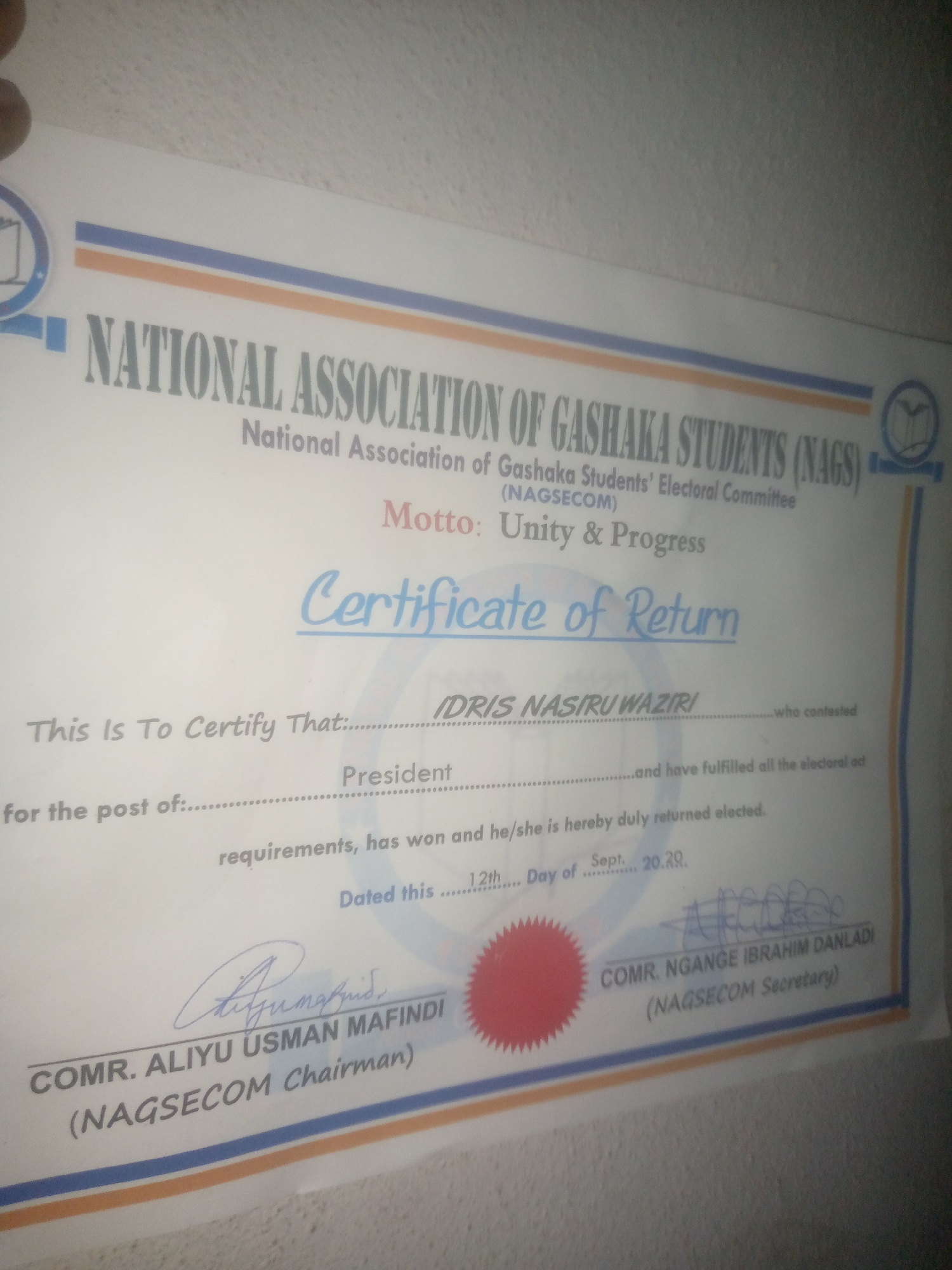 The image is a close-up, angled shot of a "Certificate of Return" issued by the National Association of Gashaka Students (NAGS). The certificate is printed on white paper with blue and orange borders. The header clearly states "NATIONAL ASSOCIATION OF GASHAKA STUDENTS (NAGS)" and "National Association of Gashaka Students' Electoral Committee (NAGSECOM)". The motto is "Unity & Progress".
The main body of the certificate reads: "This Is To Certify That: IDRIS NASIRU WAZIRI who contested for the post of: ............................................................................................................................................................................................................................................and have fulfilled all the electoral requirements, has won and he/she is hereby duly returned elected. Dated this ....... 12th......... Day of .......... Sept. .......... 20.20."
Two signatures are present at the bottom. The first is above the text "COM R. ALIYU USMAN MAFINDI (NAGSECOM Chairman)". The second signature is above the text "COM R. NGANGE IBRAHIM DANLADI (NAGSECOM Secretary)". A red seal is visible to the left of the second signature.
The image is taken indoors, likely a well-lit room, with a plain, light-colored wall in the background. The lighting casts a slight shadow and glare on the document, suggesting it is laid flat on a surface. The resolution is adequate to read most of the text, though some of the finer details of the signatures are blurred. The certificate appears to be a formal document related to an election within a student association. There are no people clearly visible in the frame beyond the signatures. The environment is neutral, without any specific location indicators.