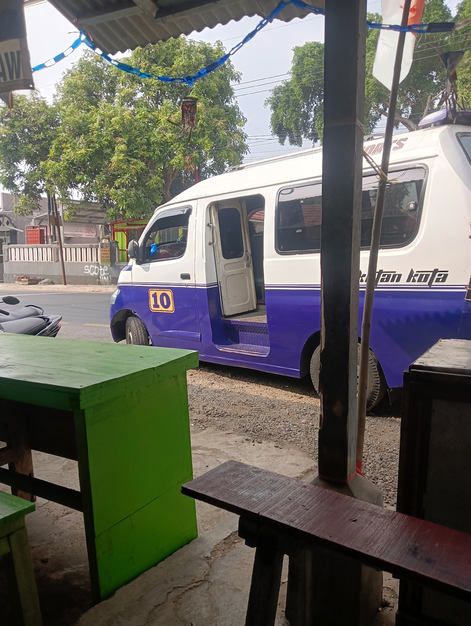 The image shows a street scene in Manggari, Indonesia. A white and purple van with the number "10" on its side is parked on a gravel lot next to a road. The van's door is open, revealing a step leading inside. A motorcycle is parked in the foreground to the left, partially obscured by a bright green table. In the background, there are trees, buildings, and a flag. The scene appears to be outside a small shop or eatery, with wooden tables and chairs visible.
