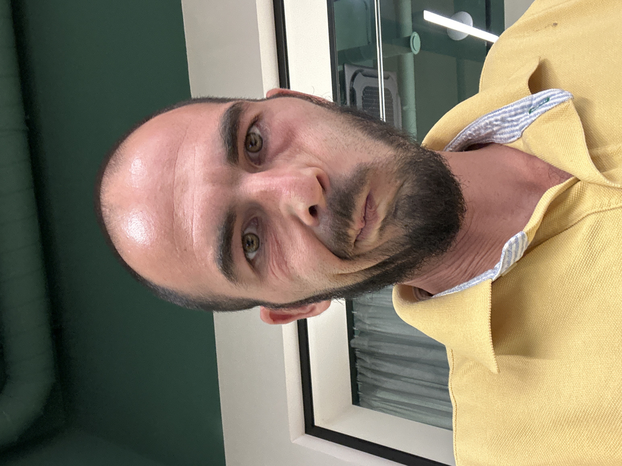 A man with a beard and receding hairline, wearing a yellow polo shirt, makes a distorted facial expression while taking a selfie. He is indoors, possibly in an office or common area, with a dark green ceiling and a light-colored wall with a window visible behind him. The location is the city of Budva, Montenegro.