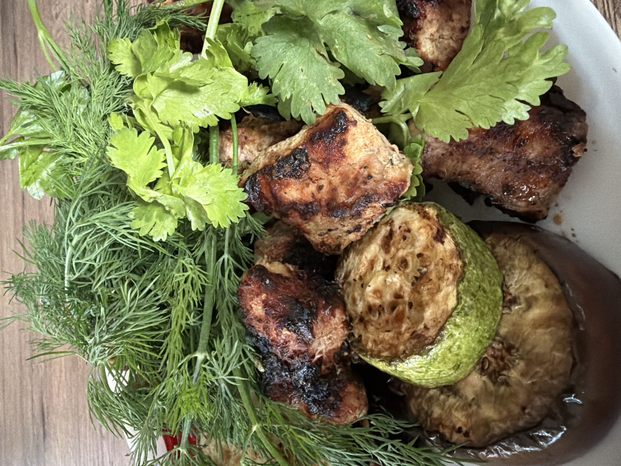 This media file is a close-up, top-down photograph of a plate of grilled food, likely a meal from Shashmurka, Russia. The primary subjects are pieces of grilled meat, possibly shashlik or kebab, alongside grilled slices of zucchini and eggplant. The meat appears to be seasoned and has char marks from grilling. The vegetables are also visibly grilled, with the zucchini having a slightly browned exterior and visible seeds, and the eggplant appearing soft and cooked through.
Surrounding the grilled items is a generous garnish of fresh green herbs. These include feathery dill and broad-leafed parsley or cilantro, adding a vibrant contrast to the cooked food. The herbs are fresh and appear to be slightly moist, suggesting they were recently washed.
The food is presented on a white plate, which is partially visible. The background shows a wooden surface, likely a table, adding a rustic element to the scene. There is no visible text in the image. The lighting suggests an indoor setting, possibly a kitchen or dining area, with soft illumination that highlights the textures and colors of the food. The overall impression is of a delicious and freshly prepared meal, typical of home-style cooking. The image does not depict any people or indicate a specific time of day or weather conditions beyond the general setting.