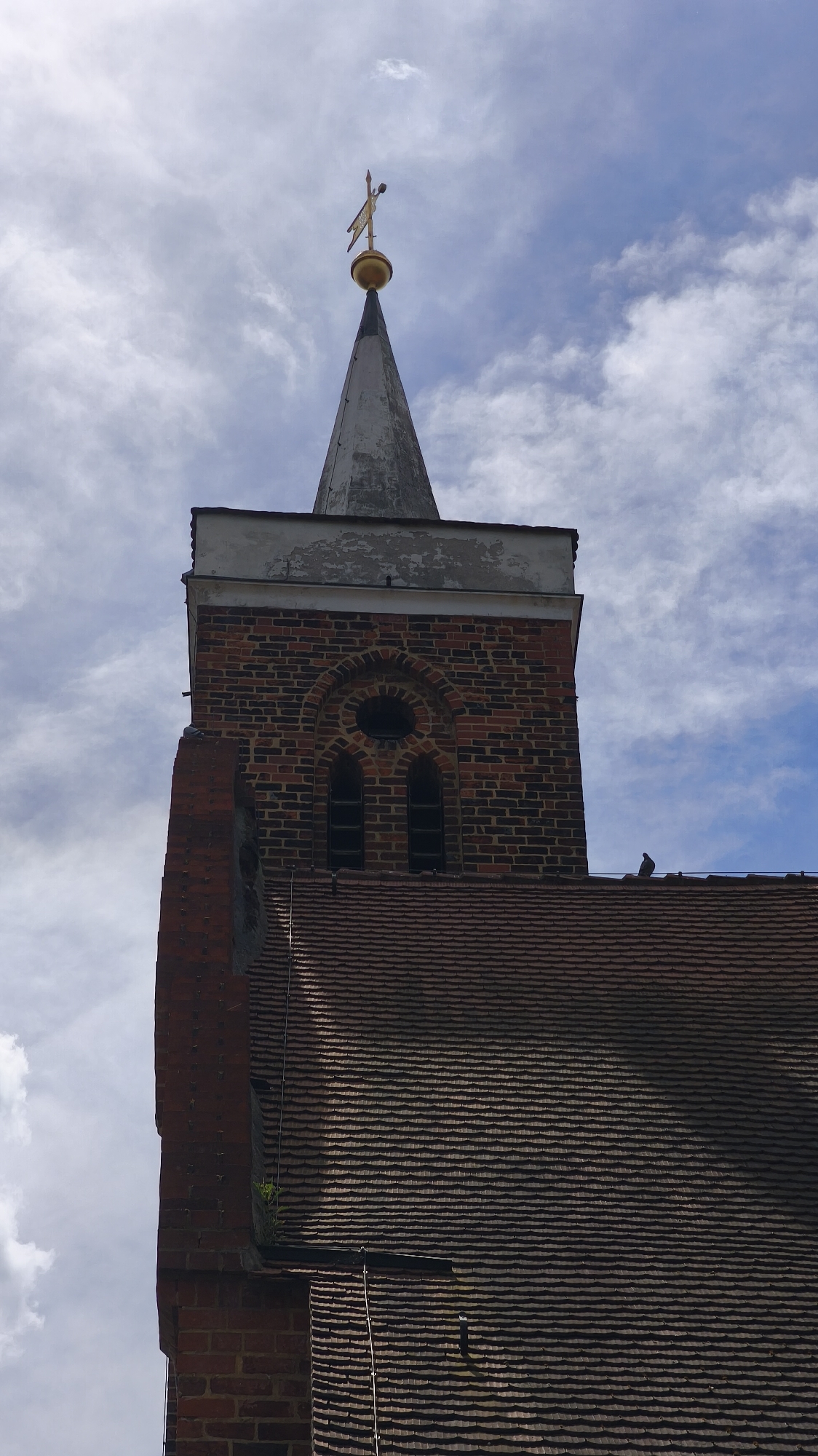 The image shows the upper section of a brick church tower with a gray spire and a gold weather vane on top. The tower has a decorative arched window above two narrow windows. The tiled roof of the church is visible below the tower. The sky is partly cloudy. The location is Unknown, Unknown.