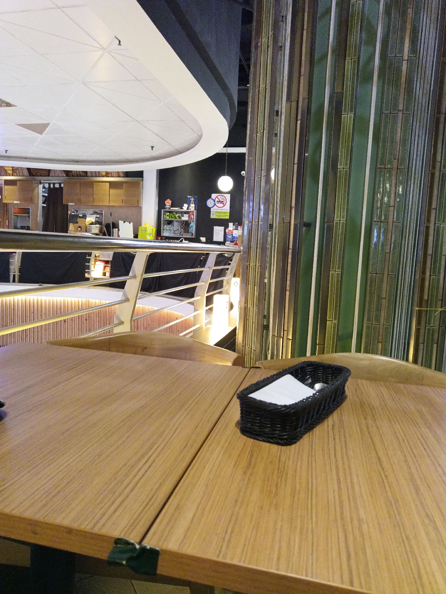 The image depicts a close-up view of a light wood grain table in a restaurant, with a black woven basket containing napkins and cutlery in the foreground. Behind the table, a section of a restaurant interior is visible, featuring a decorative bamboo-like wall on the right and a stainless steel railing and stairs in the middle ground. In the background, there are kitchen counters and cabinets, suggesting a service area. The ceiling features a modern, curved design with recessed lighting. Signs, including a "No Smoking" sign with a clock and a "Selamat Merokok" sign which translates to "Enjoy Smoking" are visible on the wall. The lighting appears to be artificial and ambient. The overall scene suggests a casual dining establishment. The specific location within Kuala Lumpur, Malaysia is not discernible from the visual elements alone.