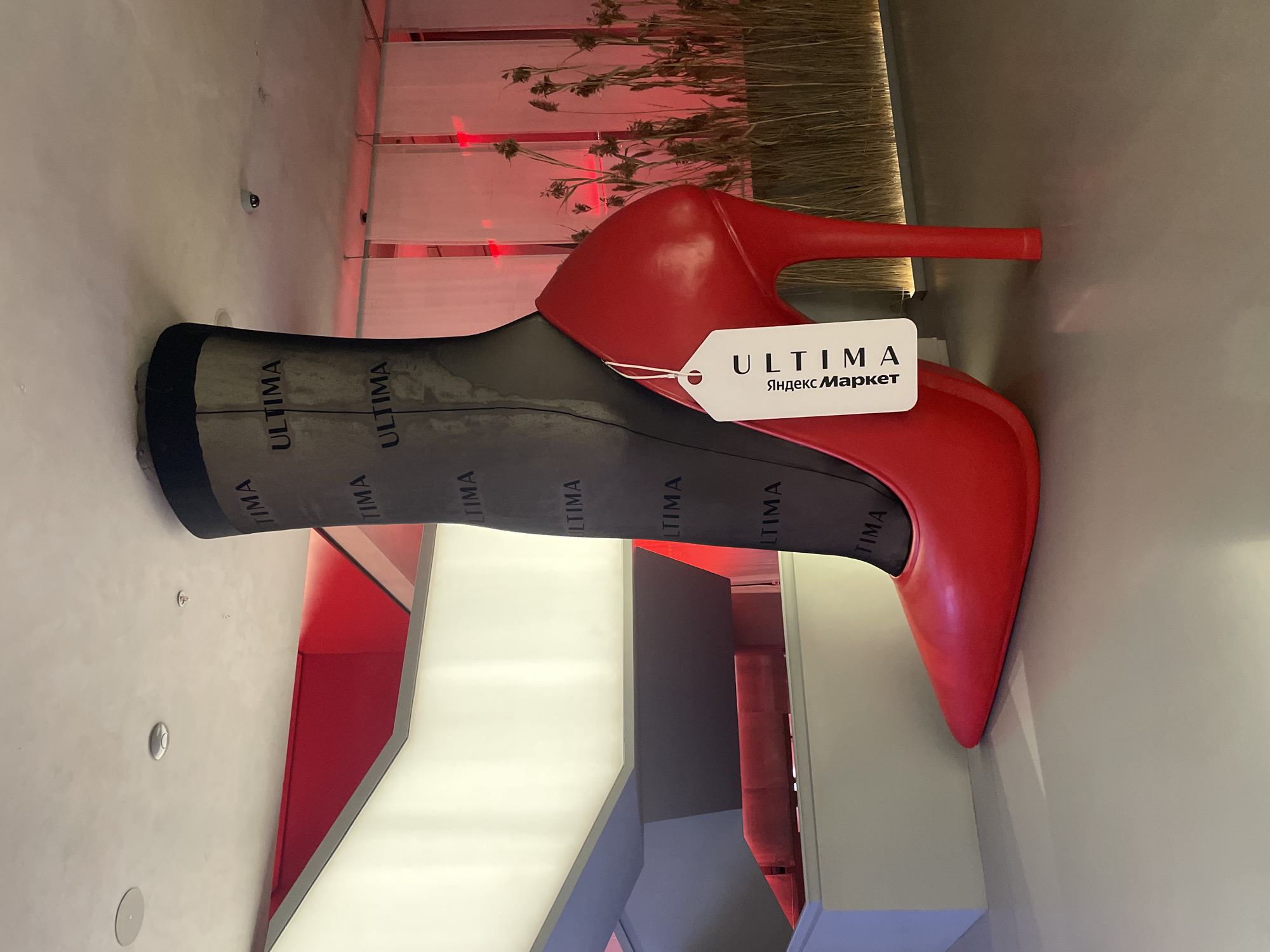 The photograph captures a large, stylized red stiletto heel with a black stocking emerging from the top. The stocking is patterned with the word "ULTIMA" repeated multiple times. A white price tag with "ULTIMA" and "Яндекс Маркет" printed on it is attached to the heel. The sculpture is situated in an indoor space with red and white architectural elements, including a tiered structure that appears to be a staircase or display. In the background, there are translucent white panels illuminated by red light and a display of dried grass or reeds. The floor is a smooth, light-colored surface. The overall impression is of a fashion-themed display or art installation, likely within a commercial or retail setting. The presence of "Яндекс Маркет" on the tag suggests a connection to the Russian e-commerce company Yandex Market. Given the context of Yandex Market, this installation is likely located in a retail space or an event associated with the brand in Saint Petersburg, Russia.
