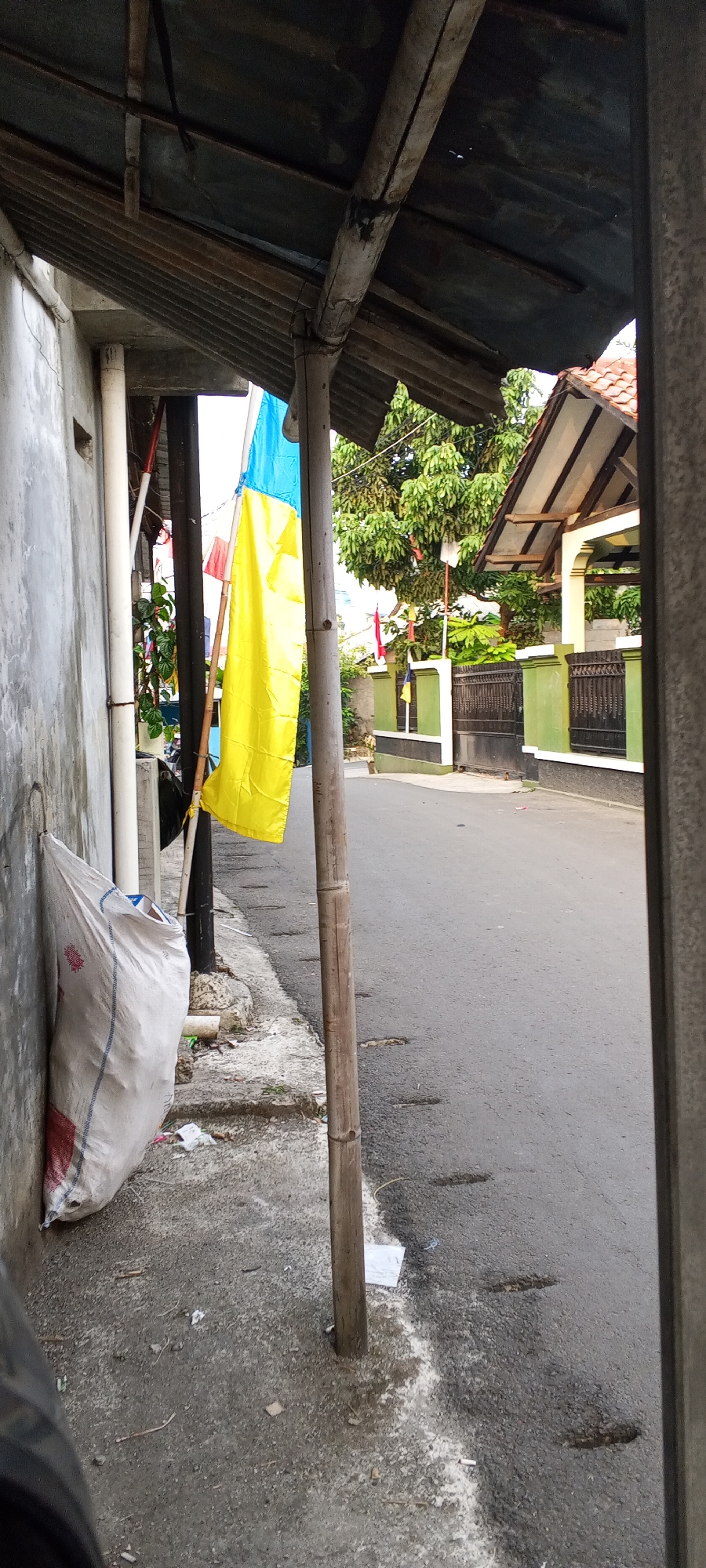 The image depicts a narrow street in Depok, Indonesia, with a flag bearing yellow and blue horizontal stripes prominently displayed on a bamboo pole. A large white sack, possibly containing trash, leans against a concrete wall on the left side of the frame. The street is paved and shows signs of wear, with several small indentations or divots. The surrounding environment includes residential buildings with tiled roofs and green fences, as well as lush green trees. A rustic overhang, constructed from corrugated metal and bamboo, provides shade above the sidewalk area. The overall scene suggests a typical urban or suburban street in Indonesia, with a focus on the flag as a potential cultural or political symbol.