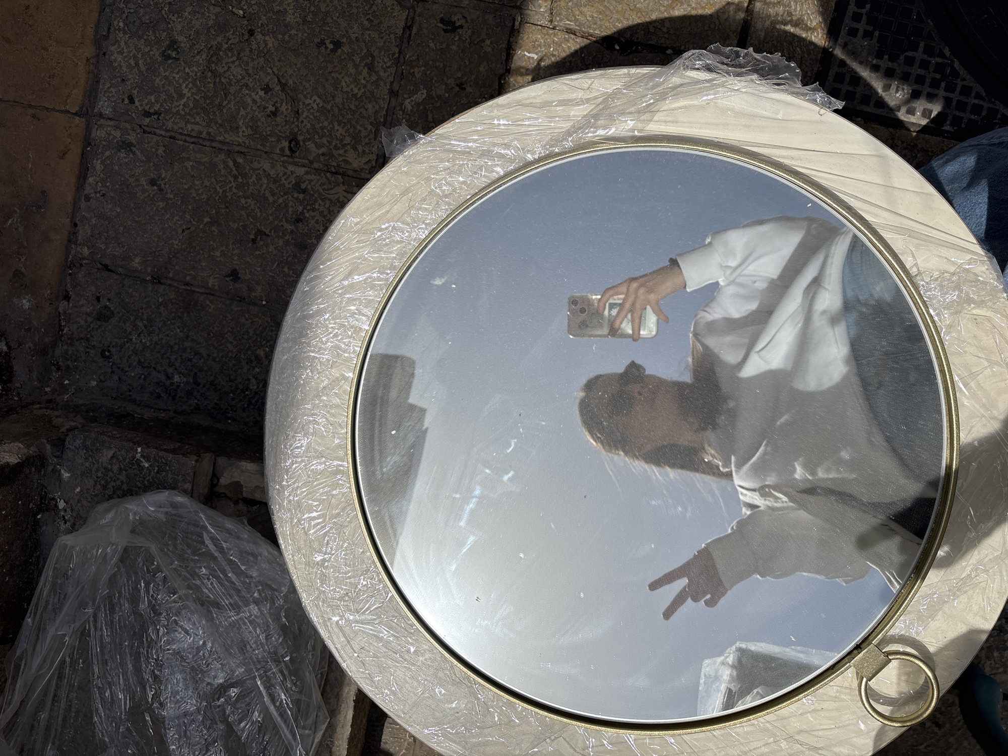 The image is a close-up, overhead shot of a round mirror with a light-colored frame, likely cream or off-white, and a thin gold accent line. The mirror is partially covered in clear plastic wrap. Reflected in the mirror is a person taking a photo of themselves with a smartphone. The person is wearing a white long-sleeved shirt and is making a peace sign with their fingers.
The mirror is placed on a textured surface, appearing to be cobblestones or weathered concrete, possibly a sidewalk or street. A crumpled plastic bag is partially visible to the lower left of the mirror, and other elements of an urban environment, such as a grated drain and possibly denim-clad legs of someone standing nearby, are also present.
Due to the presence of cobblestones and the general urban setting, the location could plausibly be Tel Aviv, Israel, but there are no specific markers confirming that.