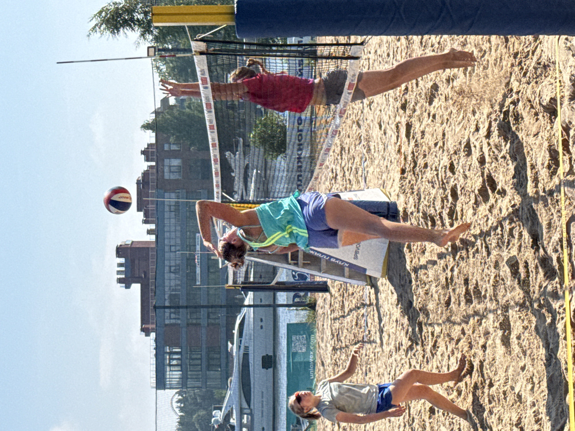 The image shows a beach volleyball game in Saint Petersburg, Russia. Three women are actively playing. One woman, wearing a teal top and blue shorts, is in mid-air, spiking the ball. Another woman, in a red top and grey shorts, is jumping to block the spike. A third woman, in a grey t-shirt and blue shorts, is positioned on the left side of the court, observing the play. The game is taking place on a sandy beach with a volleyball net set up. In the background, a large white yacht is docked on the water, and a modern building with many windows and a distinctive architectural style is visible. The sky is clear and blue.