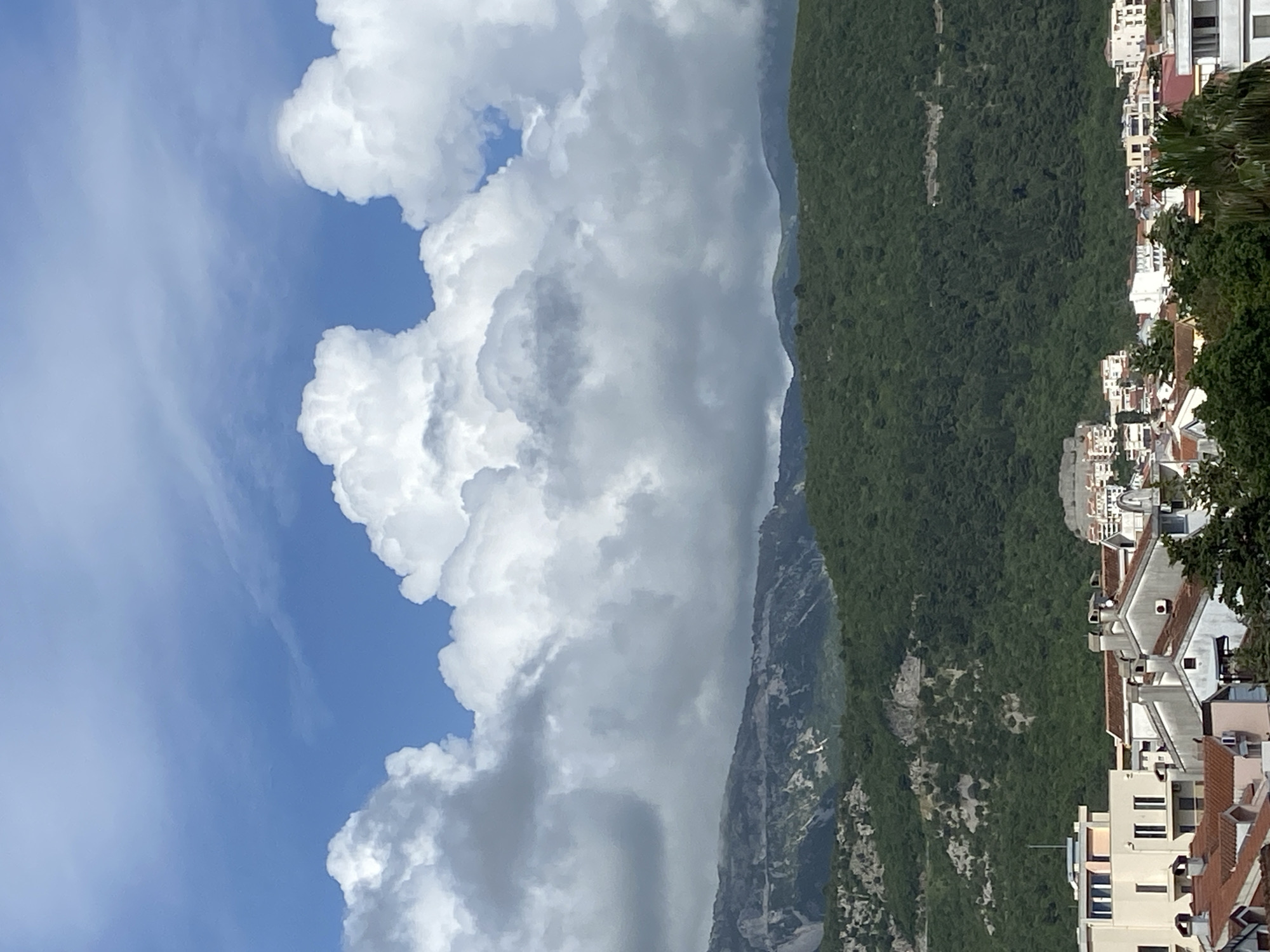 The image shows the Rock of Gibraltar from the top down, with buildings on the right and the clouds filling most of the left side. The Rock of Gibraltar is covered in green vegetation. The buildings are mostly white with red tile roofs. The clouds are a mixture of white and gray. The sky is blue.