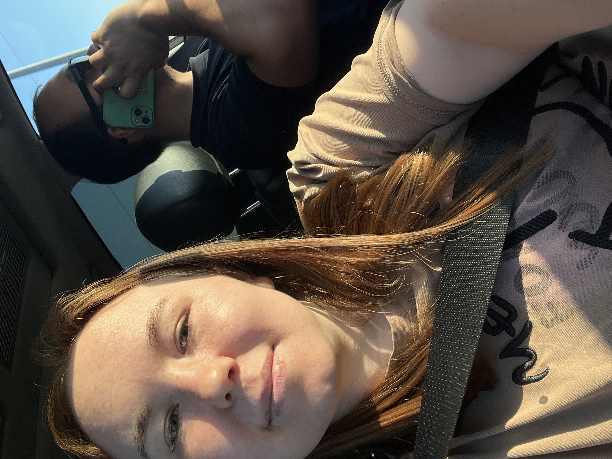 The image appears to be a selfie taken inside a car. A woman with red hair is in the foreground, wearing a seatbelt and a light brown t-shirt. Behind her, partially out of focus, is a man holding a phone with a green case to his face. He is wearing a black shirt. The interior of the car is visible, including the headrest of the driver's seat. The location is not discernible from the image, and there is no evidence to suggest that the image was taken in the city of Chaikovsky, Russia.