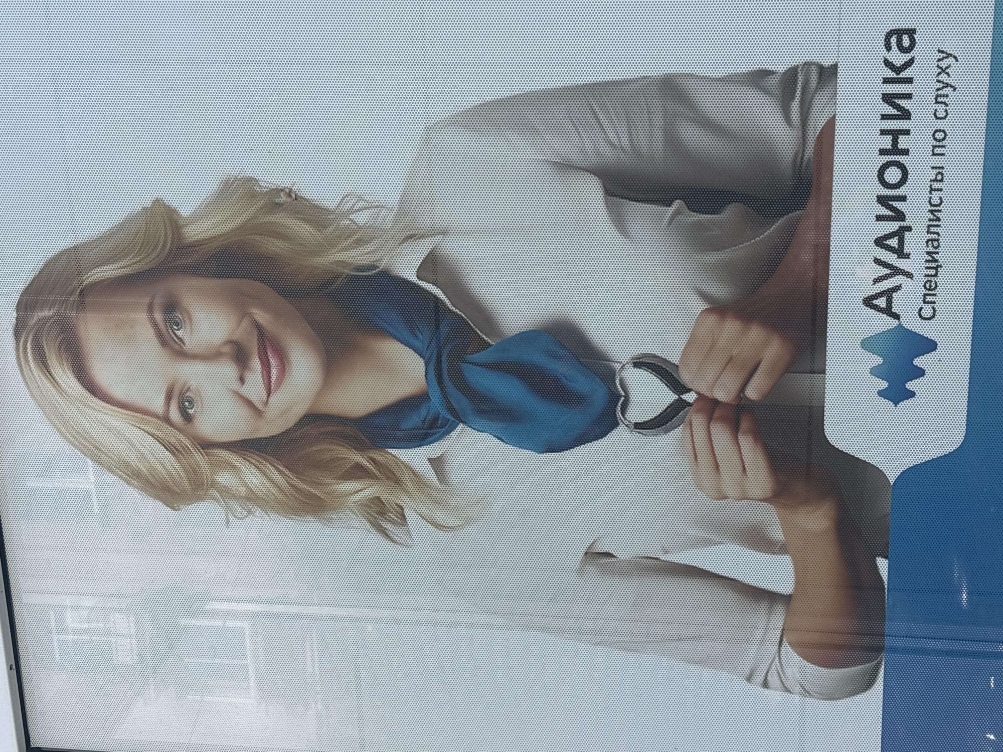 The image shows a large advertisement for "Аудионика Специалисты по слуху" (Audionika Specialists for Hearing). The advertisement features a woman with blonde hair wearing a white shirt and a blue scarf. She is smiling and holding a heart-shaped stethoscope to her chest. The background of the advertisement is a light blue with a white dotted pattern, and it appears to be a digital display or a perforated banner. The advertisement is located outdoors, as indicated by the presence of a building visible in the upper left corner and a blue reflective surface at the bottom, which could be a window or a display case. The context suggests this is a commercial advertisement likely found on a street or in a public area within Novosibirsk, Russia, given the Cyrillic text on the advertisement.