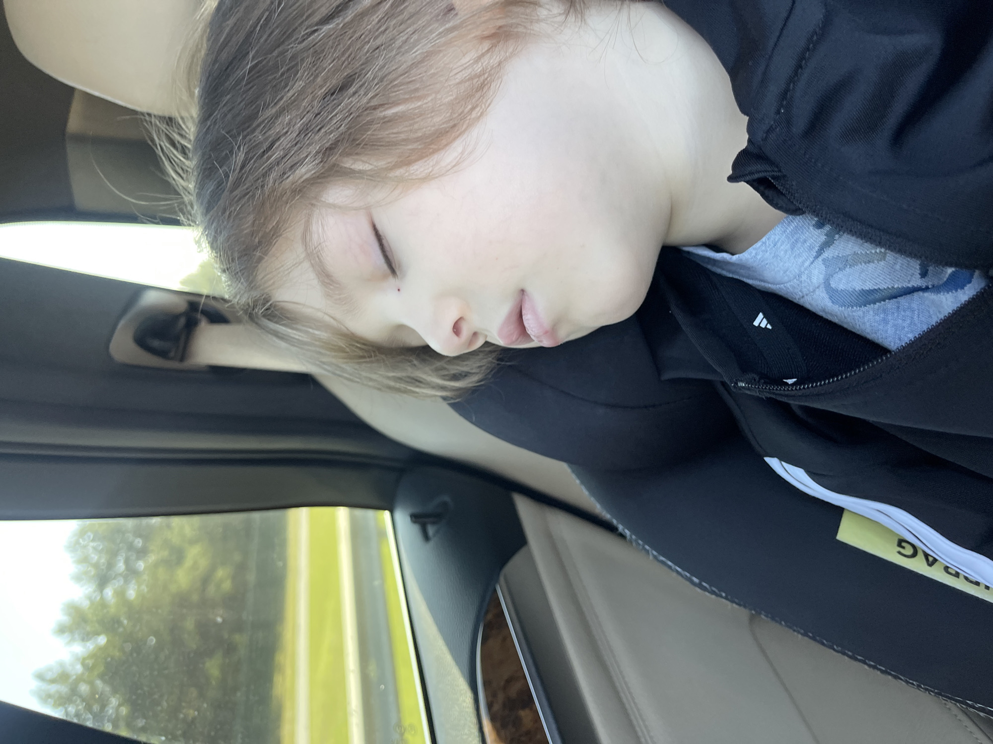 A child is sleeping in a car seat. The child's eyes are closed, and their head is resting on the side of the car seat. The child is wearing a black jacket over a grey t-shirt. A yellow tag that says "AIRBAG" is visible sticking out of the car seat. Through the window, trees and a road are visible. The interior of the car appears to be made of beige leather. The image was taken inside a vehicle, which is not in Tallinn, Estonia.