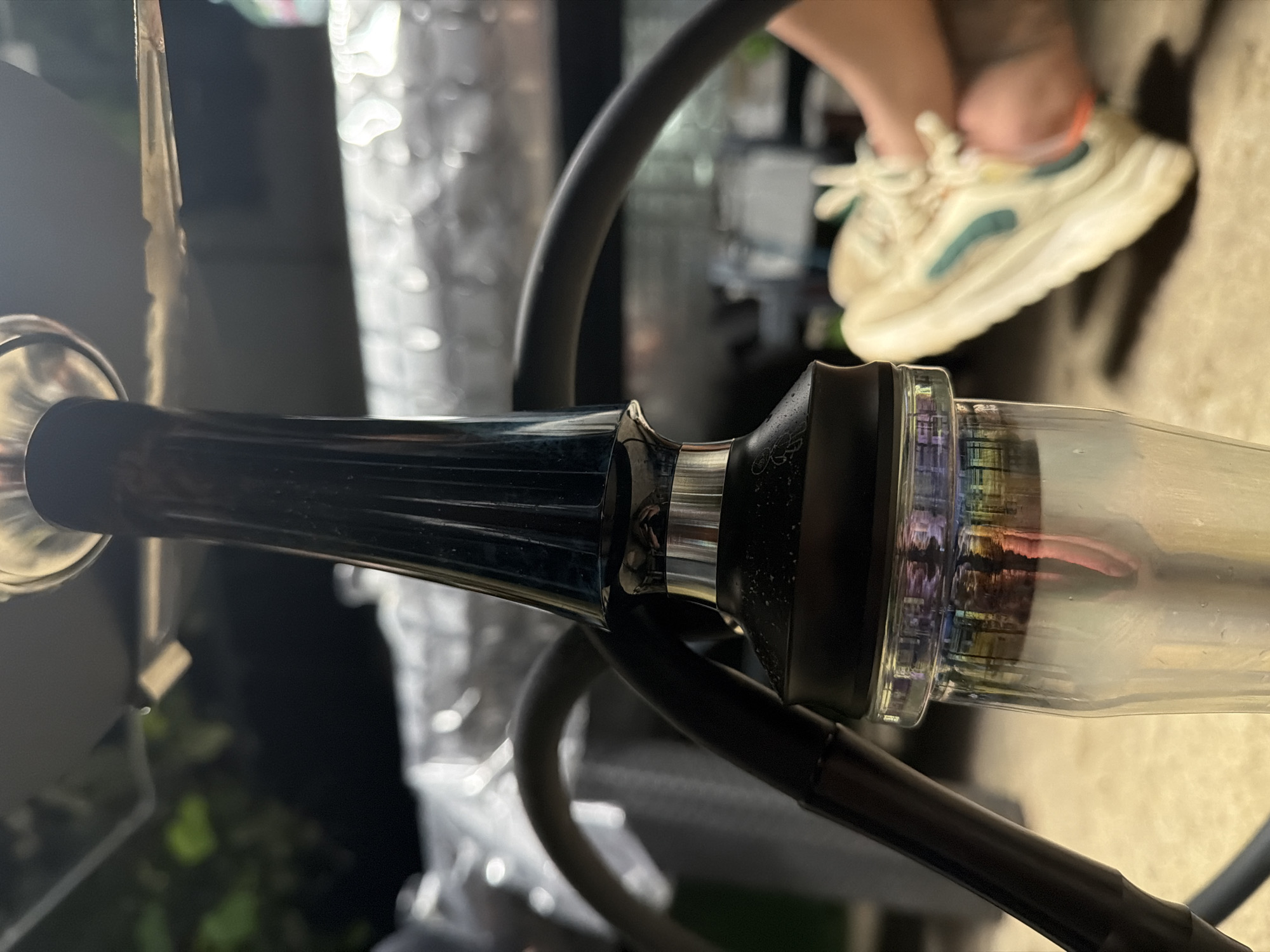 This image captures a close-up, low-light scene featuring a hookah in the city of Svetovrachene, Bulgaria. The central subject is a dark-colored hookah, likely dark blue or black, with a ribbed stem and a prominent silver band at its lower section. A black, ribbed hose is attached to the hookah's mid-section. The hookah rests on a light-colored, textured ground surface, possibly concrete or pavers. Its clear glass base contains water, exhibiting colorful internal reflections in shades of pink, yellow, blue, and green, suggesting the presence of an internal LED light or external colored illumination.
In the blurred background, a person's right foot is visible, wearing a cream-colored athletic sneaker with distinct teal and orange accents. The foot appears to be resting on the ground, indicating the person is likely seated or reclined nearby. Further in the background, out of focus, are dark areas consistent with outdoor foliage or darkness, and lighter, reflective surfaces that may be part of a table or other outdoor furniture, possibly covered with a white plastic sheet or similar material. The overall lighting suggests an evening or nighttime setting. No explicit actions are depicted beyond the static arrangement of objects and the resting foot.