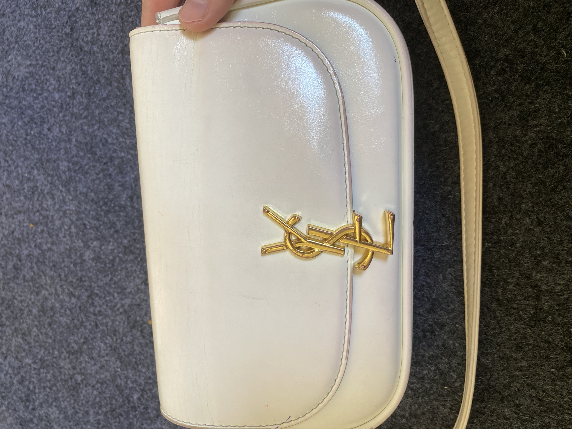 A white, rectangular handbag with a gold interlocking "YSL" logo is centered in the frame. The handbag has a curved flap closure and a thin, light-colored strap. The material appears to be leather and shows some minor scuff marks and signs of wear. The handbag rests on a dark gray, textured surface, possibly a carpet or rug. The background is out of focus, but the surface appears consistent with the foreground. There is a human hand visible on the right side of the image, lightly touching the edge of the handbag. No other people or significant environmental details are visible. The location in Arbagish, Uzbekistan is not discernible from the image.