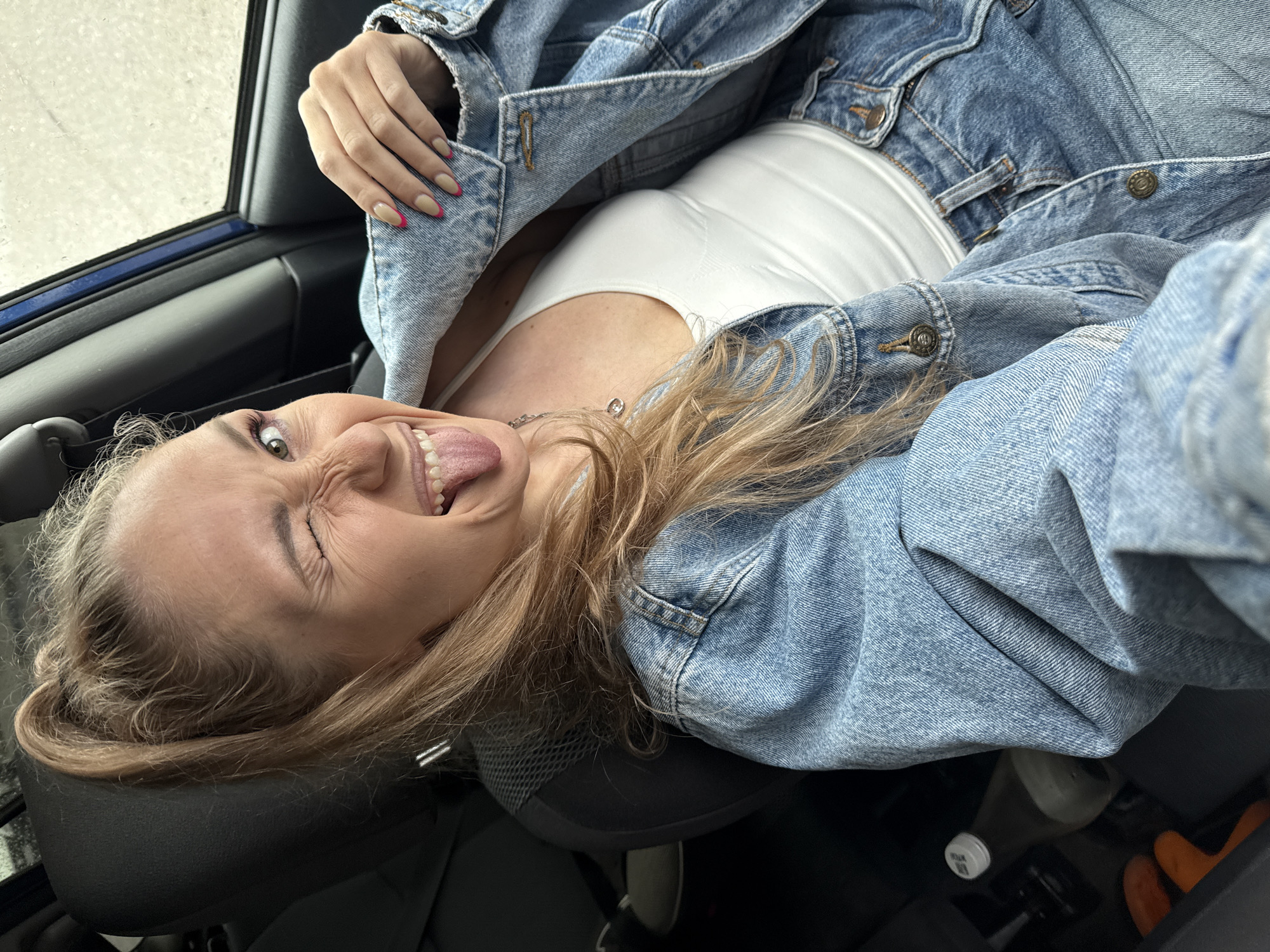 The image shows a young woman inside a car. She is leaning back in the driver's seat, wearing a white tank top and a blue denim jacket. She is sticking her tongue out and winking with one eye. Part of the car's dashboard and window is visible. The photo appears to have been taken inside a vehicle, but no location information confirms it is in Chaikovsky, Russia.