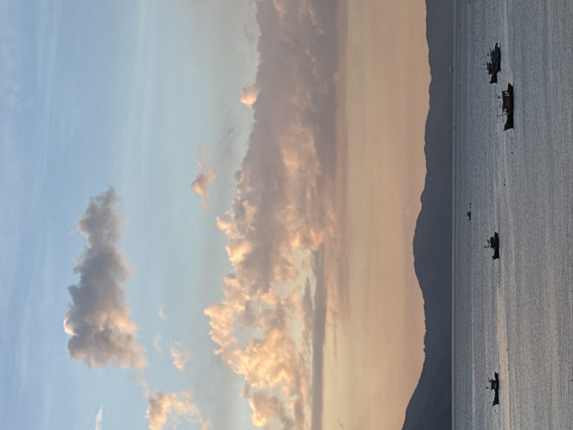 A wide shot of the ocean and sky at sunset. The sky transitions from a pale blue at the top to hues of orange and pink near the horizon. Clouds are scattered across the sky, some tinged with pink from the setting sun. A dark, silhouetted mountain range forms the backdrop against the horizon. In the foreground, the calm ocean reflects the colors of the sky. Several small fishing boats are scattered across the water, their silhouettes dark against the water. The scene evokes a peaceful and serene atmosphere at the end of the day. This image was taken at sunset over the ocean near Sơn Trà Peninsula, Đà Nẵng, Vietnam.
