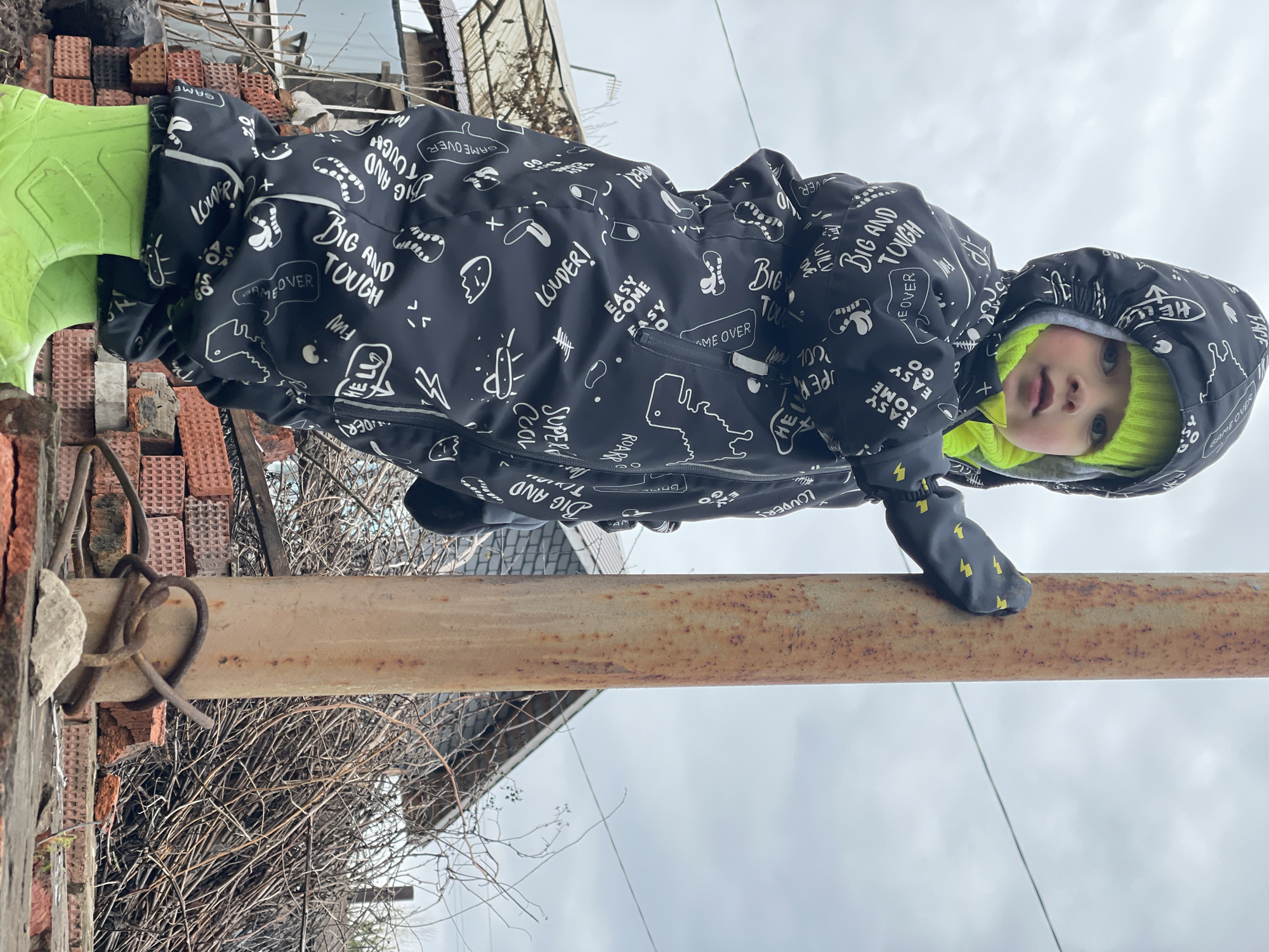 A young child in a black and white printed snowsuit and bright green boots and hat is balancing on a rusty metal bar outdoors. The child is looking at the camera with their right hand gripping the bar. To the left of the child are stacks of red bricks and bare branches. In the background, there is a building and an overcast sky. The location is suggested as Zav'yalovo, Russia.