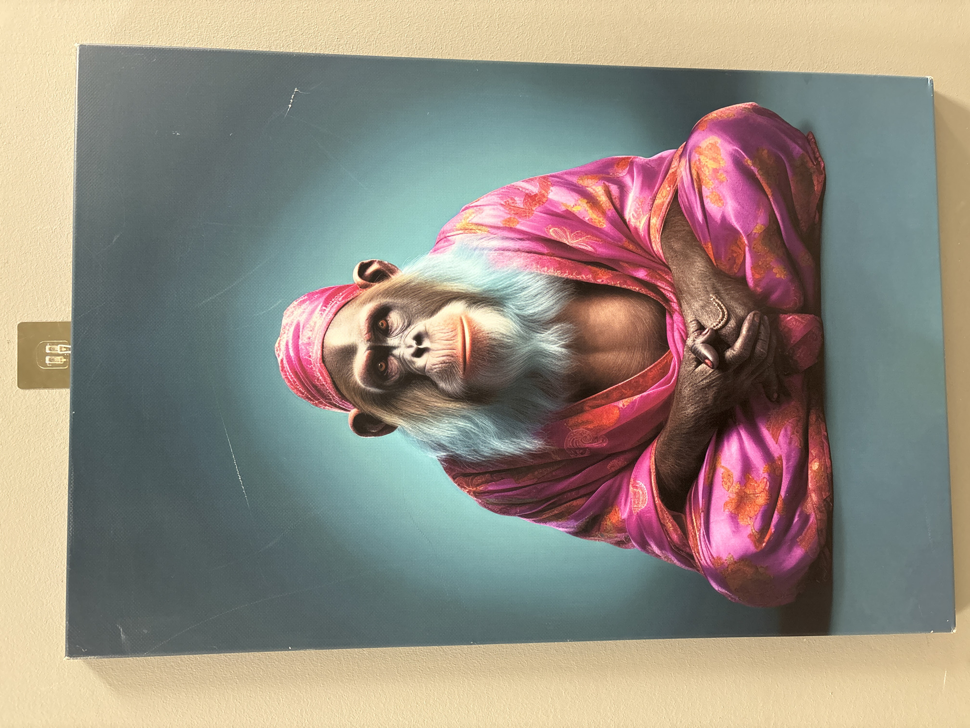 This is a close-up, slightly tilted photograph of a canvas print featuring a stylized portrait of a chimpanzee. The chimpanzee is depicted as if sitting in a meditative pose, wearing a pink and orange patterned silk robe and a matching pink headscarf. The chimpanzee has light blue fur around its face and neck, contrasting with its dark fur. Its hands are clasped in its lap. The background of the print is a gradient of teal to a lighter, bluish-green, with a spotlight effect originating from the upper right. The canvas appears to be mounted on a wall, with the wall visible in the top and bottom sections of the image. There are some faint white scratches on the surface of the canvas print. A small, grey adhesive mounting strip is visible on the left edge of the canvas. The lighting suggests an indoor setting, with no indication of time of day or weather. The emotion conveyed by the chimpanzee is one of calmness or contemplation. There is no visible text in the image.