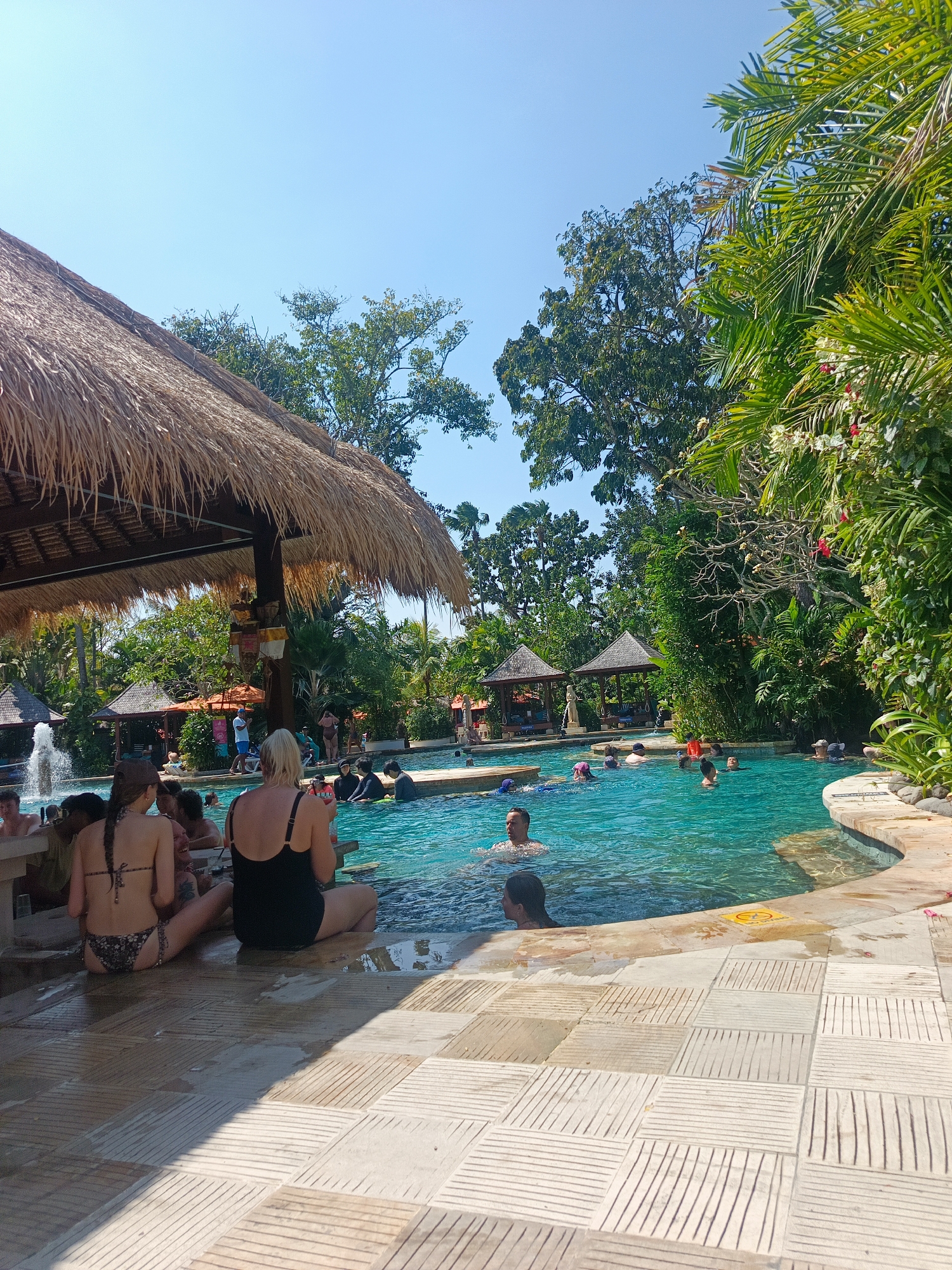 In Kuta, Indonesia, a tropical outdoor swimming pool area is depicted under a clear blue sky. A thatched-roof structure, resembling a bar or shaded seating area, extends into the frame from the left. Several people are visible, some seated on the tiled poolside in the foreground, facing away from the camera. One individual wears a black swimsuit, while another is in a patterned bikini. Numerous individuals are in the turquoise swimming pool, some swimming, others socializing, and a few appear to be children.
The pool itself has a curved edge with ornate stone coping. In the background, lush green trees and tropical foliage surround the pool area, with a few small, traditional-looking pavilions with pitched roofs scattered amongst the greenery. A fountain is visible on the far left, with water spraying upwards. Sunlight casts distinct shadows on the tiled patio in the foreground. The overall atmosphere suggests a vibrant, recreational setting, likely a resort or hotel amenity in the popular tourist destination of Kuta.