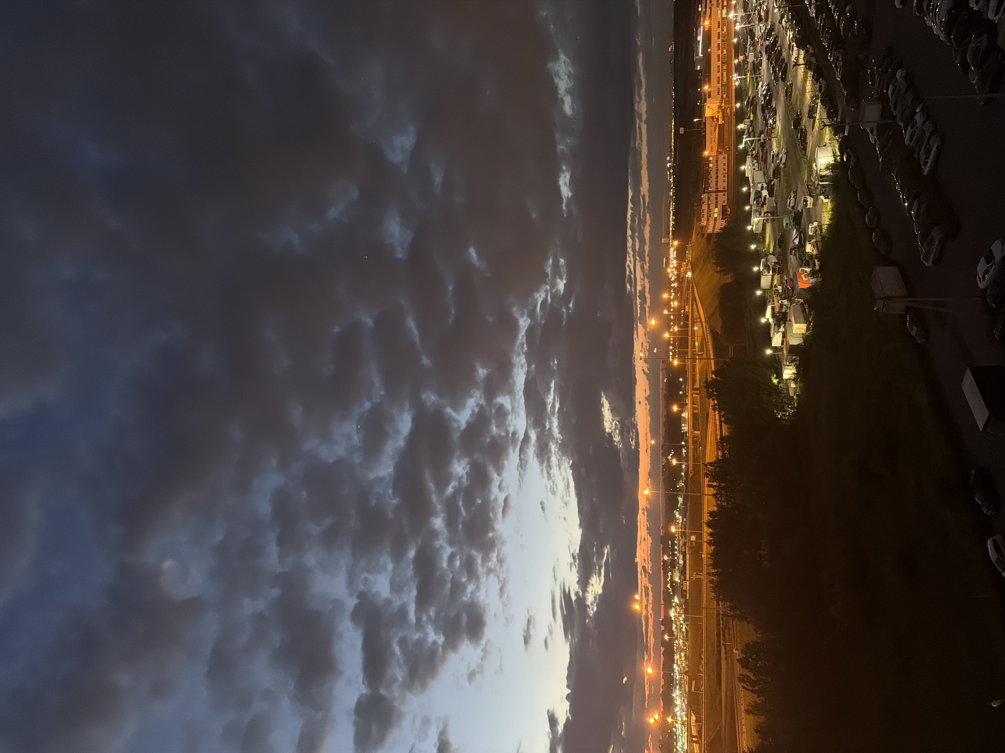 The image depicts an aerial view of a large parking lot filled with numerous cars, trucks, and trailers, illuminated by overhead lights. In the background, buildings with glowing windows and streetlights are visible, suggesting an urban or industrial area. The scene is set against a twilight sky with dramatic clouds, colored with hues of orange and deep blue, indicating either sunrise or sunset. The foreground shows the dark silhouette of trees and a few parked cars.