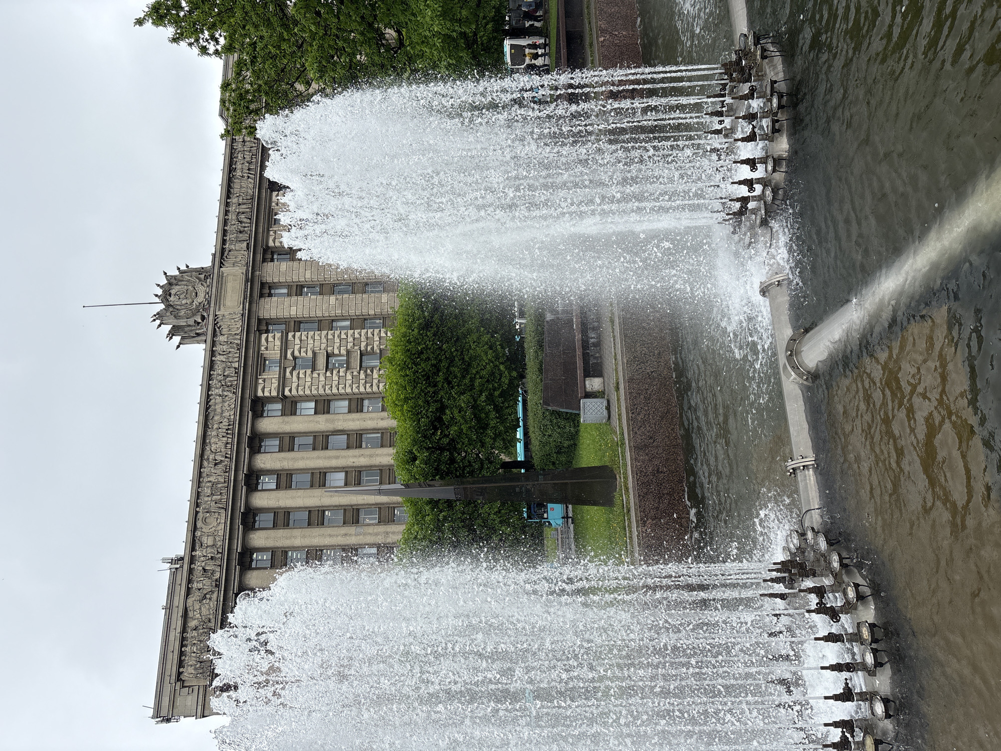 The image shows a fountain in Saint Petersburg, Russia, with jets of water shooting upwards. In the background, there is a large, grey building with multiple windows and ornate carvings on its facade. The fountain is made of metal pipes and valves, with water spraying from multiple nozzles. The environment appears to be overcast. In the background, there is a blue bus behind the fountain. No people are clearly visible in the image.