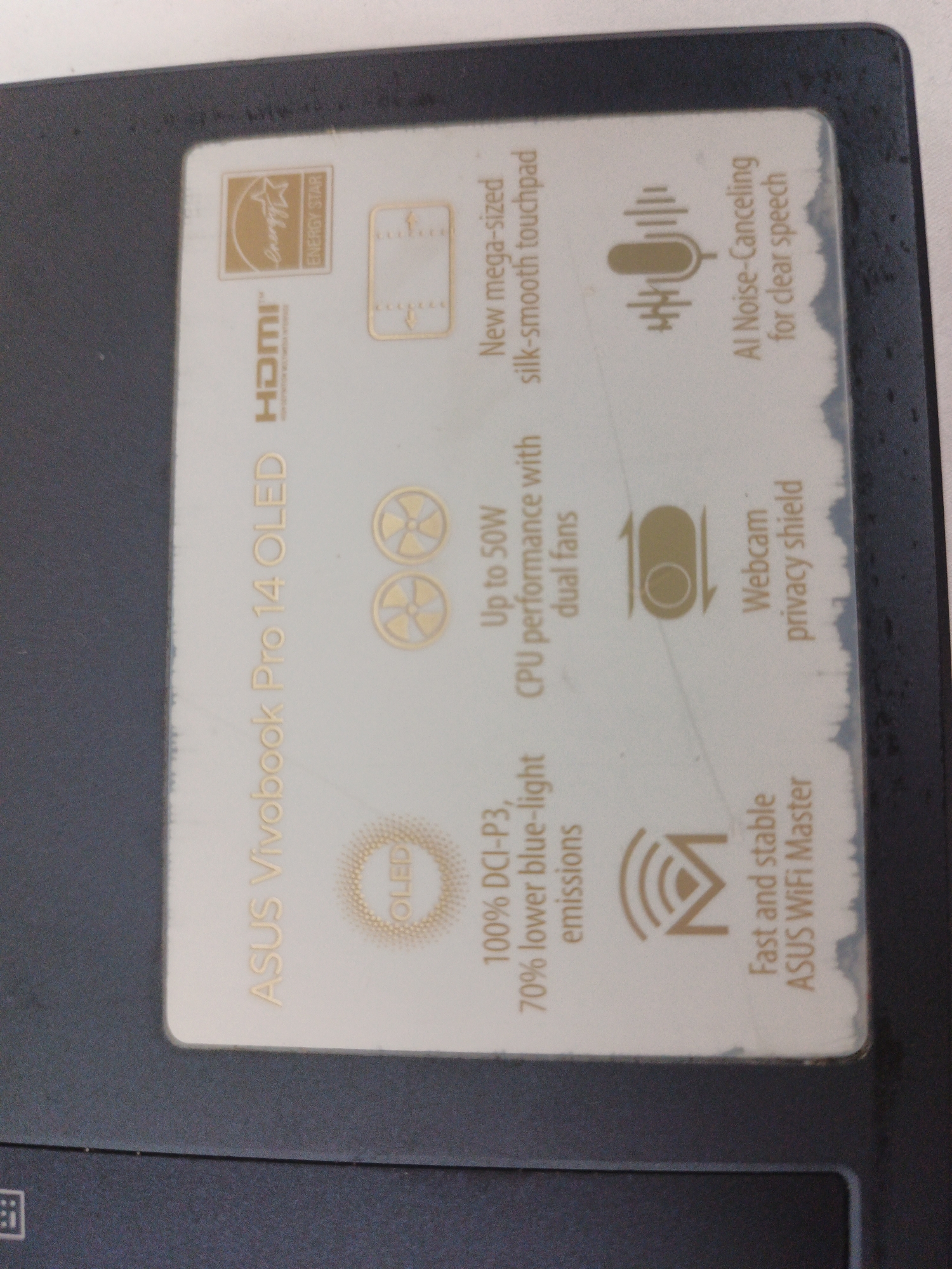 This is a close-up, eye-level, indoor, horizontal shot of a white sticker with gold text and icons adhered to a dark grey surface. The sticker appears to be on a laptop lid. From top to bottom, the sticker contains an "ENERGY STAR" logo, an HDMI logo, the text "ASUS Vivobook Pro 14 OLED", a graphic of two spinning fans, text describing "Up to 50W CPU performance with dual fans", a microphone icon with text "AI Noise-Canceling for clear speech", a circular icon with arrows and text "Webcam privacy shield", a circular graphic labeled "OLED", text stating "100% DCI-P3, 70% lower blue-light emissions", a WiFi symbol, and text "Fast and stable ASUS WiFi Master". The sticker is slightly worn and scuffed, with some damage to the edges. The background is out of focus. There are no people or discernible environmental features beyond the laptop.