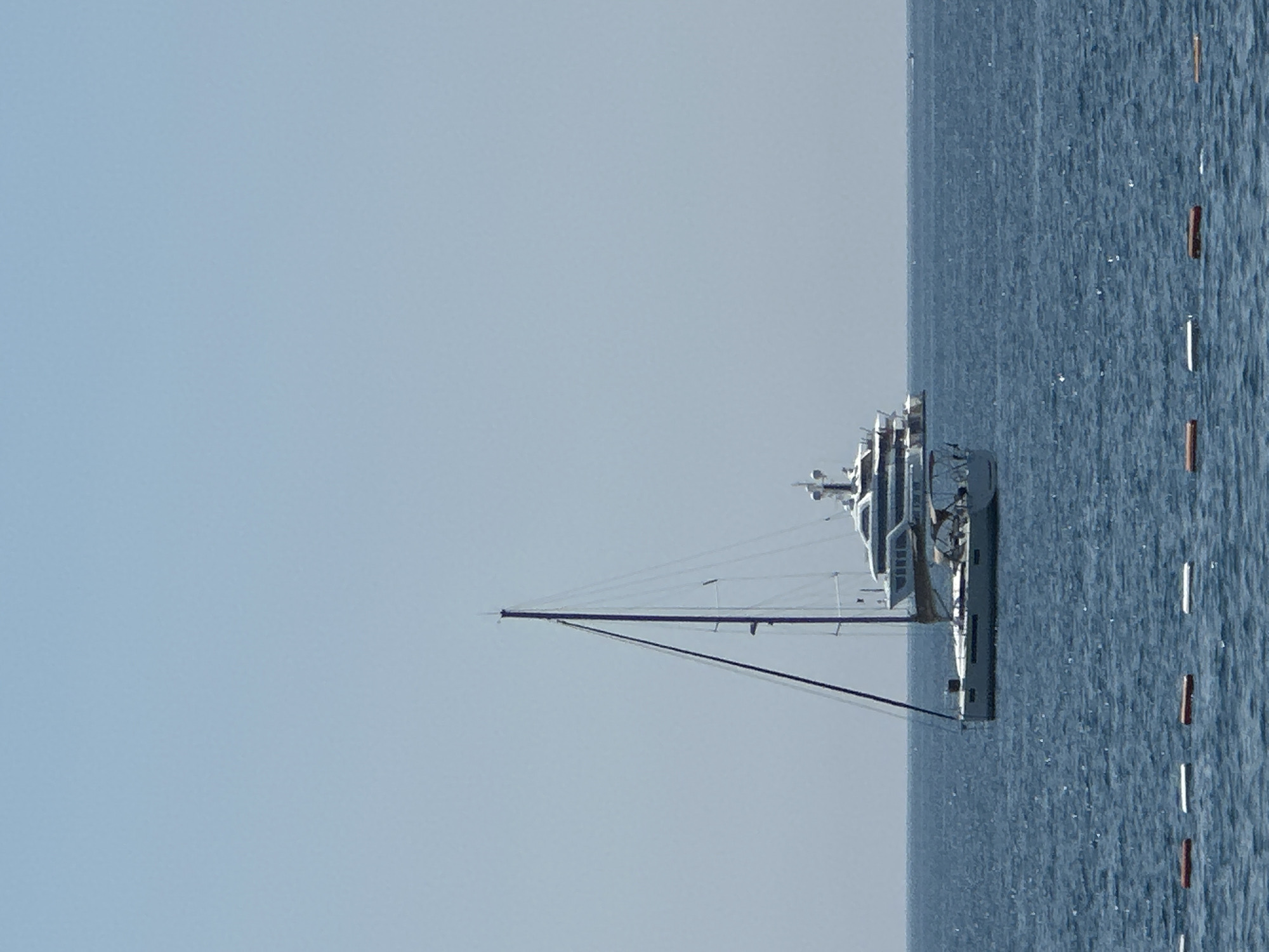 A serene scene unfolds on a clear day, likely in the morning or early afternoon given the bright, even lighting. A large, modern yacht is anchored in the calm, deep blue waters of Budva, Montenegro. The yacht has multiple decks and appears to be white and grey, with visible windows and railings. A tall mast from a sailboat is prominent in the foreground, extending diagonally across the frame and pointing towards the sky. The water is a rich blue, with small ripples indicating a gentle breeze. Along the right edge of the image, a series of buoys or markers, some white and some orange, are spaced out in a line. The horizon is clearly defined, separating the sea from a pale, cloudless sky. There are no people visible in the frame, contributing to the tranquil and peaceful atmosphere of the scene. The overall impression is one of a leisurely day at sea, with the impressive yacht and sailboat suggesting a setting for recreation or travel.