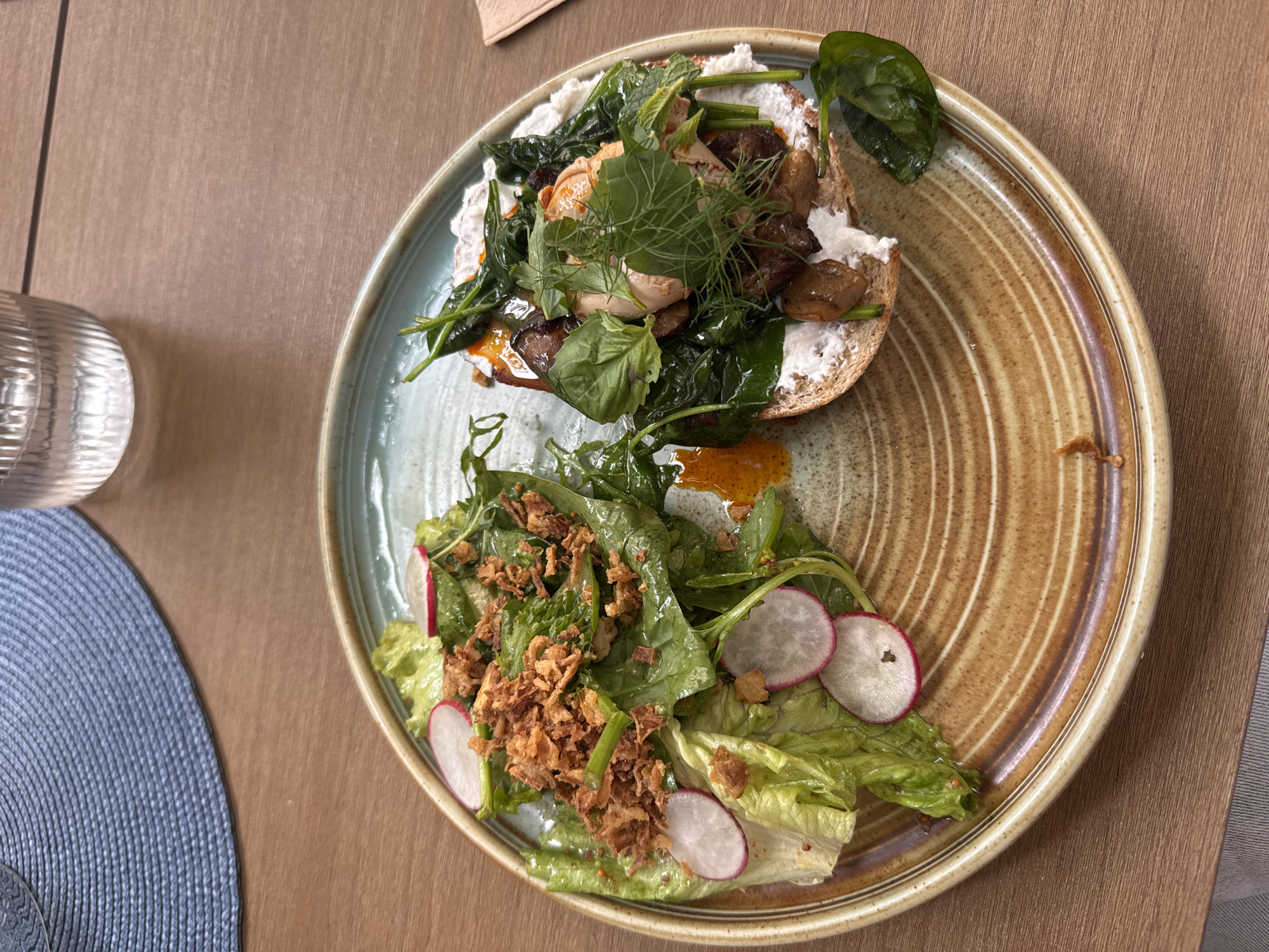 An overhead shot captures a light brown, rustic ceramic plate on a wooden table. The plate is divided into two halves, each with a different food item. On the left, a vibrant green salad is piled high, featuring crisp lettuce leaves and thin slices of radishes. Scattered atop the salad are golden-brown fried onions, adding a textural contrast. A light orange dressing is visible, drizzled over the greens. On the right side of the plate, a slice of whole-wheat bread serves as the base for an open-faced sandwich. This sandwich is generously topped with a white spread, likely cream cheese or a similar soft cheese. Dark, sauteed mushrooms are interspersed with fresh green spinach leaves and delicate sprigs of dill and parsley. The dish is artfully arranged, showcasing the fresh ingredients. In the upper left corner of the image, a blue woven placemat is partially visible. To the right of the placemat, a clear, ribbed glass is filled with a light-colored beverage, likely water. The background consists of the wooden table surface, with a hint of a grey napkin or tablecloth in the lower left corner.