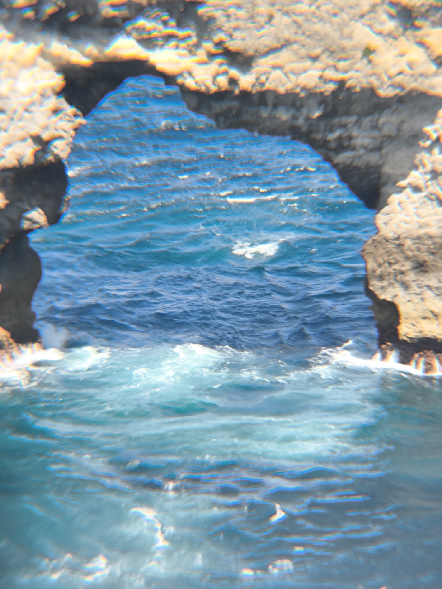 The image shows a rocky archway overlooking the ocean in Nusa Penida, Indonesia. The arch is made of light-colored rock and frames a view of the blue ocean with waves. The water appears turbulent, with some whitecaps visible. The foreground is blurred.