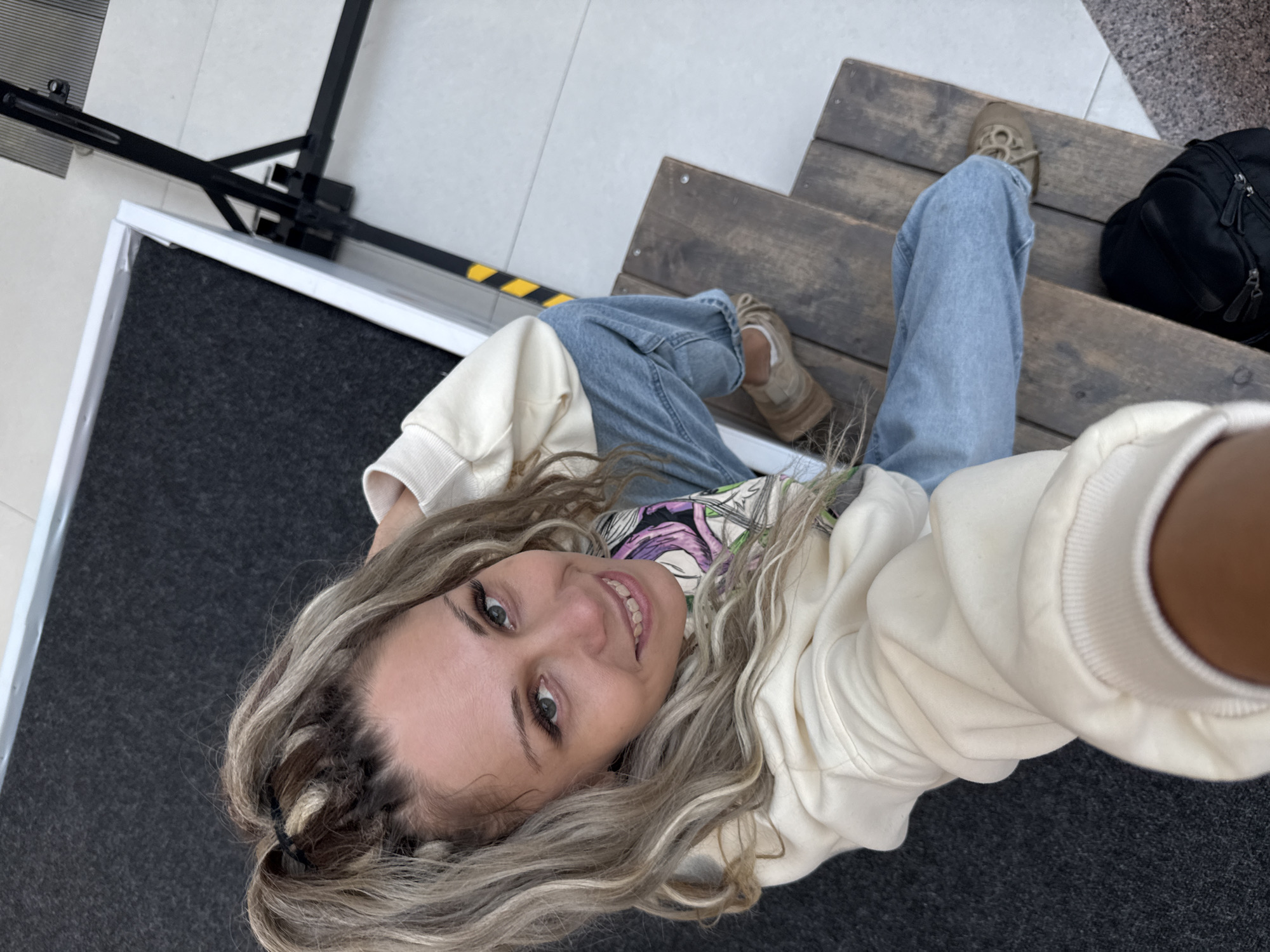 A high-angle, medium shot captures a woman from the chest up, seated on wooden steps. Her hair is a blend of blonde and darker tones, styled in two pigtails with braids. She wears a light-colored hoodie and light-wash, wide-leg jeans, paired with beige sneakers. The woman is looking up and smiling towards the camera, with her right hand extended, suggesting she is taking a selfie. The background consists of dark grey carpeted surfaces and a white tiled floor, with a metal ramp and black supports visible to the right. The scene appears to be an indoor public space, possibly a ramp leading into a building or an area designed for accessibility. There is no indication of the location being in the city of Rusanovo, Russia.