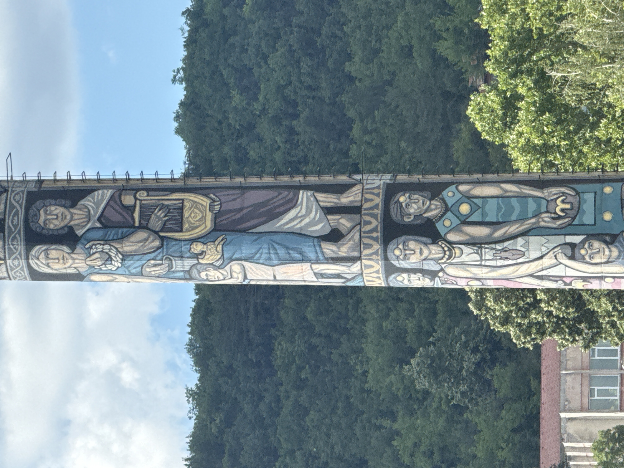 A tall, cylindrical monument adorned with a mural depicting figures in classical attire is situated in a landscape with a dense forest-covered hill in the background. The mural features several individuals, including a woman holding a laurel wreath and a man playing a lyre in the upper section. Below them, more figures are depicted, some with elaborate jewelry and holding objects. The monument's surface is textured, and a series of horizontal metal elements are attached to its side. At the base of the monument, a building with a reddish-brown roof and white-framed windows is partially visible, nestled amongst lush green trees. The sky above is blue with scattered white clouds.