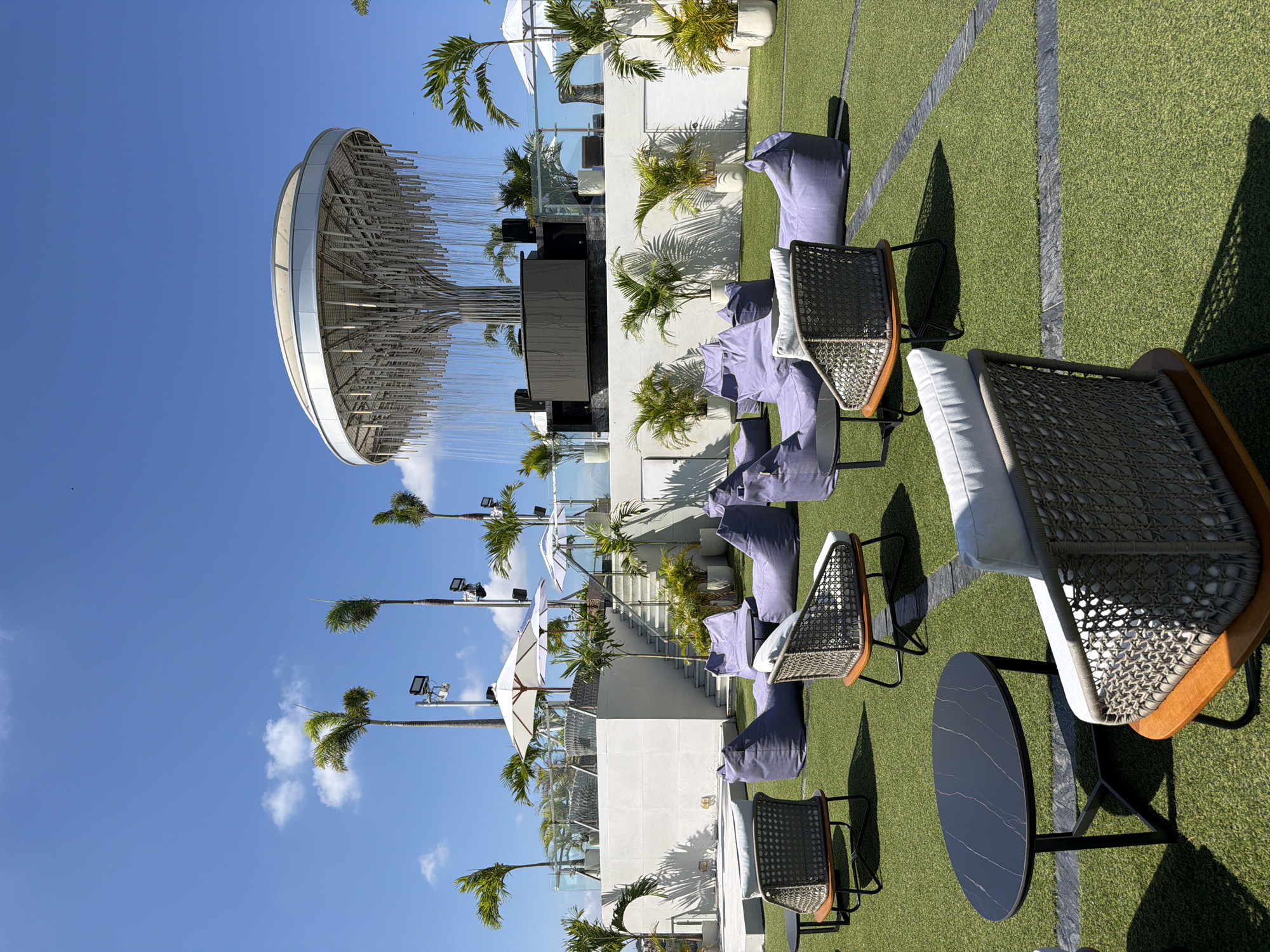 The image captures a vibrant, sunny outdoor rooftop lounge or entertainment area, suggestive of an upscale resort or hotel in North Kuta, Indonesia. The ground is covered with meticulously maintained artificial green turf, patterned with dark linear sections. Modern outdoor furniture is arranged across this space, including wicker-style chairs with light-colored cushions and several large, plush purple bean bags. Small, round black tables with subtle white marbling patterns are interspersed among the seating arrangements.
In the background, pristine white multi-level structures, possibly terraces or buildings, are visible, decorated with numerous potted palm trees that cast distinct shadows across the surfaces. Tall, slender palm trees with green fronds punctuate the bright blue sky, some featuring attached spotlights or cameras. White patio umbrellas are strategically placed for shade. A set of stairs can also be seen, leading up to an elevated section.
A prominent feature in the upper left of the scene is a large, circular, suspended art installation or decorative lighting fixture. It boasts a sleek white outer rim encasing an intricate structure of numerous thin, reflective rods or wires. The clear, vibrant blue sky is dotted with a few soft white clouds. The strong, defined shadows indicate bright daylight, likely mid-morning to late afternoon. No people are visible, but the setup clearly indicates a space designed for leisure, social gatherings, or events. There is no visible text in the image.