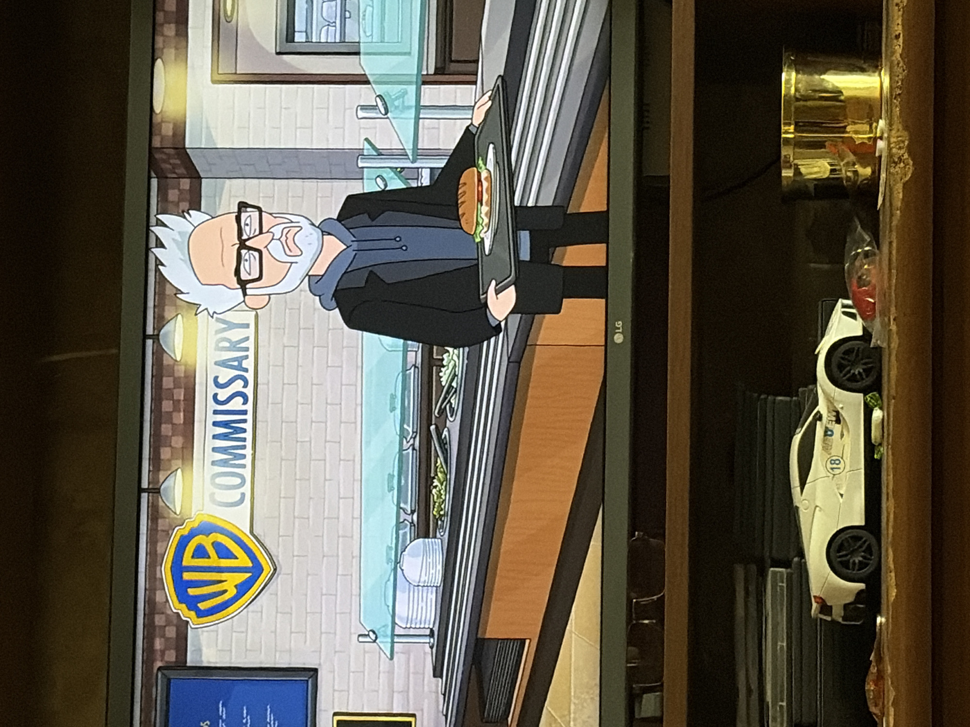 A cartoon of a man with white hair, a beard, and glasses stands in a commissary holding a tray with a sandwich. The commissary has a buffet line with food behind a glass partition and the WB logo is displayed on the wall. Below the television screen is a shelf with a white toy car, some DVDs, and a gold container. The scene is set in the city of Unknown, Unknown.