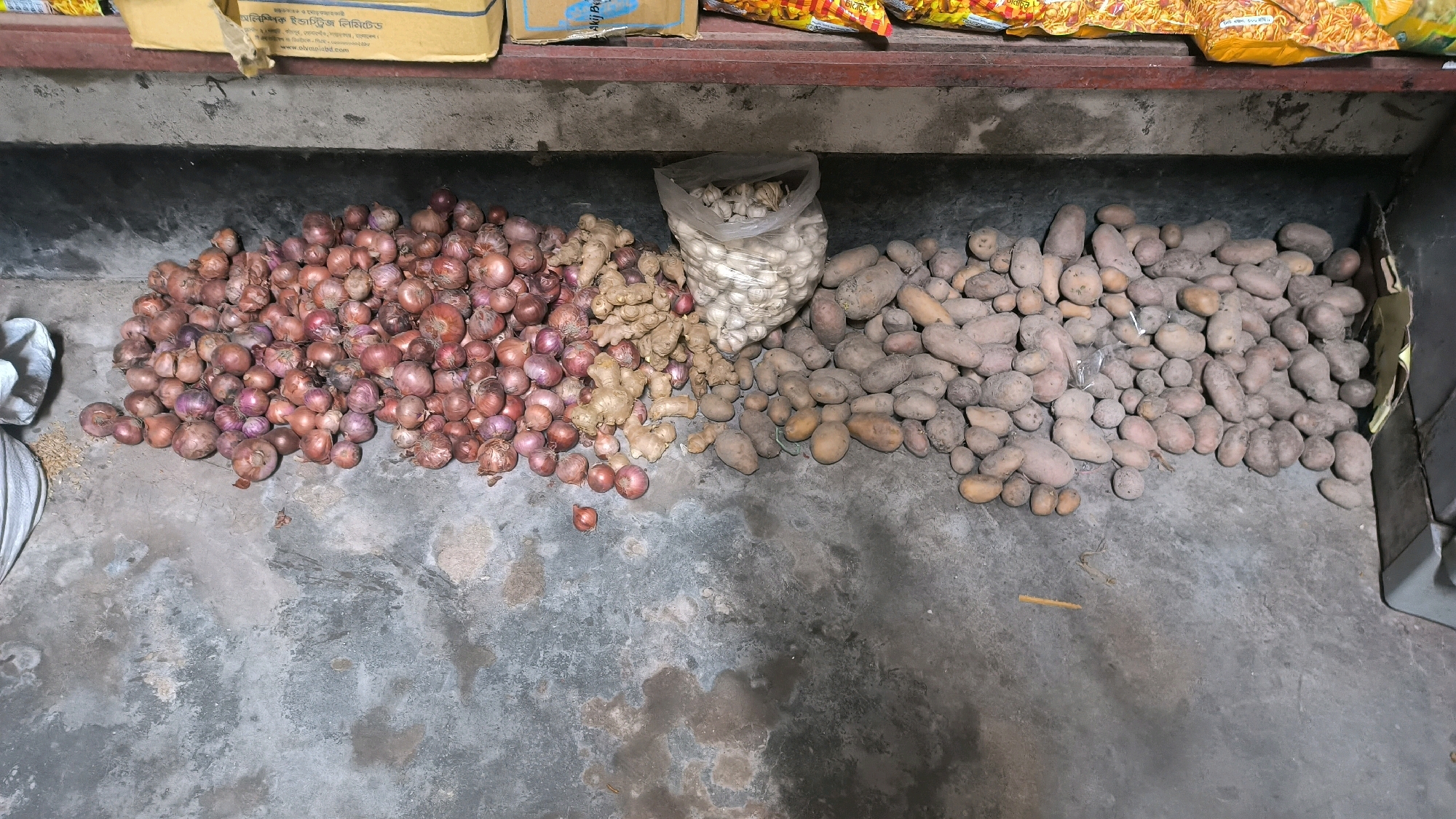 The image shows a ground-level, overhead view of various produce items scattered on a concrete floor, likely in a storage area. To the left, a large pile of red onions is visible. Adjacent to the onions, in the center, is a pile of ginger. A plastic bag filled with garlic is placed behind the ginger. To the right of the garlic bag, a large collection of potatoes of varying sizes is spread across the floor. A white sack and some loose grains are partially visible on the far left. In the background, the edge of a shelf or counter is visible, with some boxes and packets of food items resting on it. The overall scene depicts a disorganized storage of fresh produce. The location is specified as Begumganj Upazila, Bangladesh.