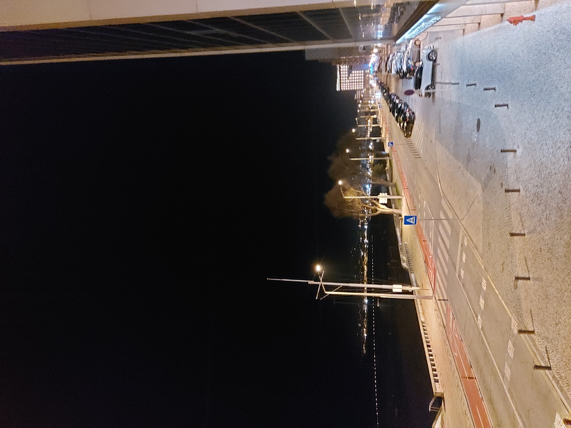 Here's a forensic description of the image:
**Location:** Figueira da Foz, Portugal, at night. The scene appears to be from an elevated position, possibly a building overlooking the waterfront.
**Environment:** A clear night sky contrasts against the lights of the city. Street lighting illuminates a wide walkway/sidewalk and street.
**Objects:**
* A row of parked cars and motorcycles line the street near a brightly lit building.
* Regularly spaced bollards separate the walkway from the street.
* Streetlights illuminate the path.
* A zebra crossing is visible on the street.
* The waterway is marked by a vertical line of lights, potentially a reflection or lighting along a bridge or bank.
**Subjects/Actions:** The scene is static, devoid of visible people or actions.
**Interactions:** None are observable in the still image.