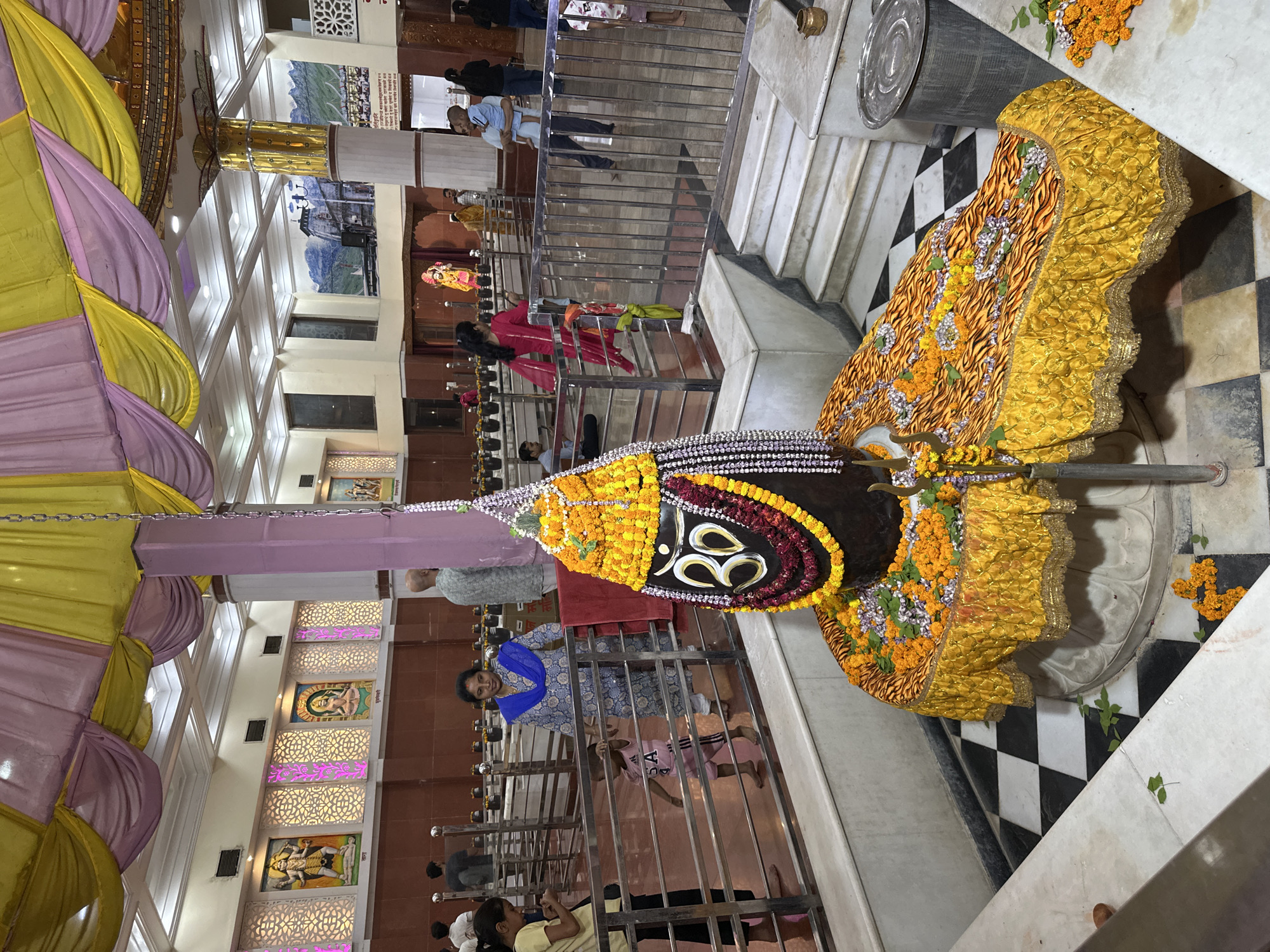 The image shows a Hindu temple scene. Centrally featured is a Shiva Lingam, decorated with garlands of yellow flowers and a large Om symbol. The Lingam is draped in golden fabric and positioned on a checkered black and white tiled floor.
People are present, some standing behind a metal railing. A woman in a blue patterned outfit stands near the railing, while others are visible in the background, including individuals on stairs. The temple interior is adorned with paintings and decorative lighting.
The location is Varanasi, India.