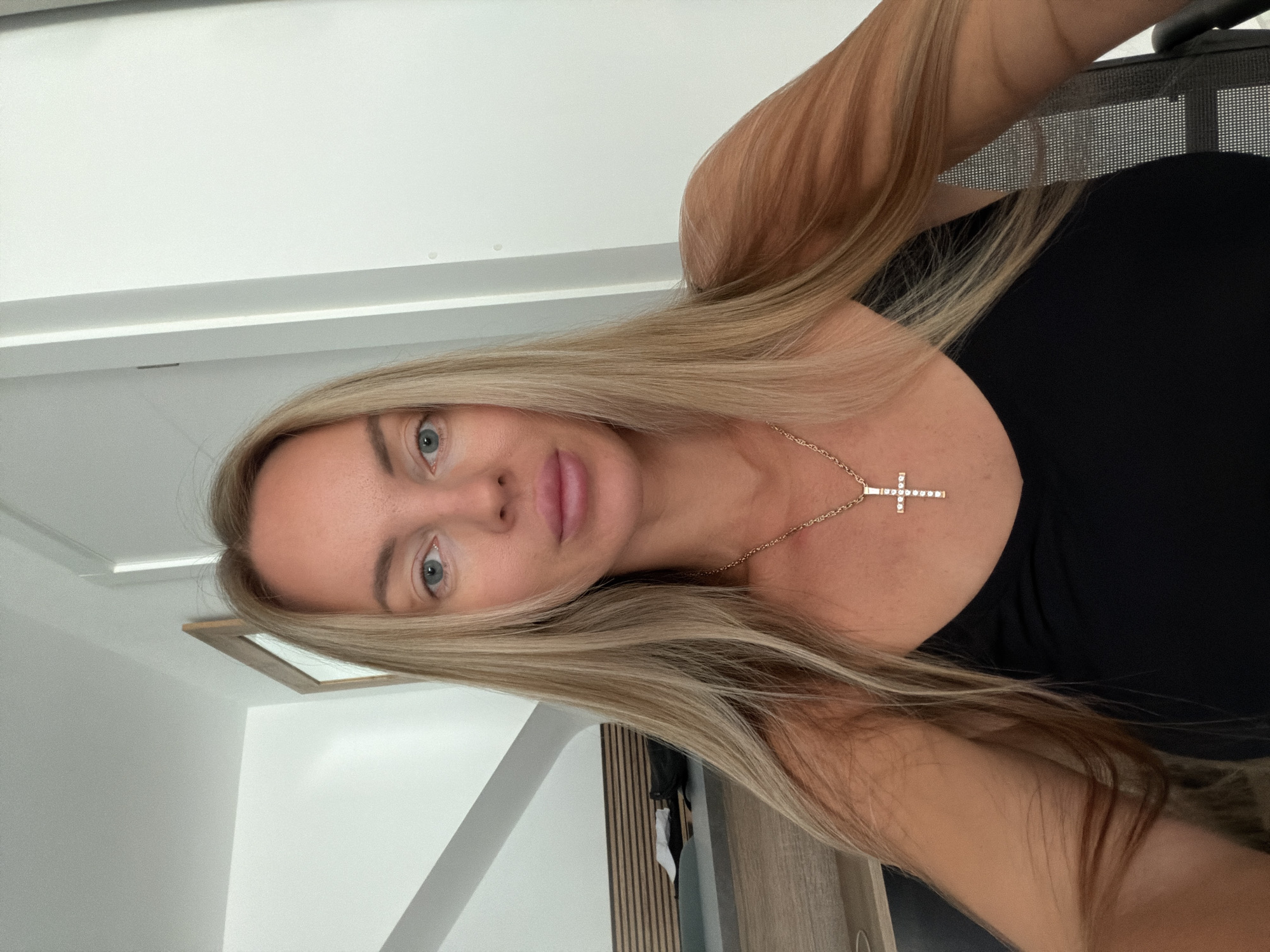 A woman with blonde hair and blue eyes is captured in a selfie. She is wearing a black tank top and a gold necklace with a cross pendant. The background appears to be an indoor setting, possibly a room in Finestrat, Spain, with a white wall and a glimpse of what might be a door or closet. Part of a wooden dresser or desk with a shelf is visible to the left. The lighting is bright, suggesting daytime.
