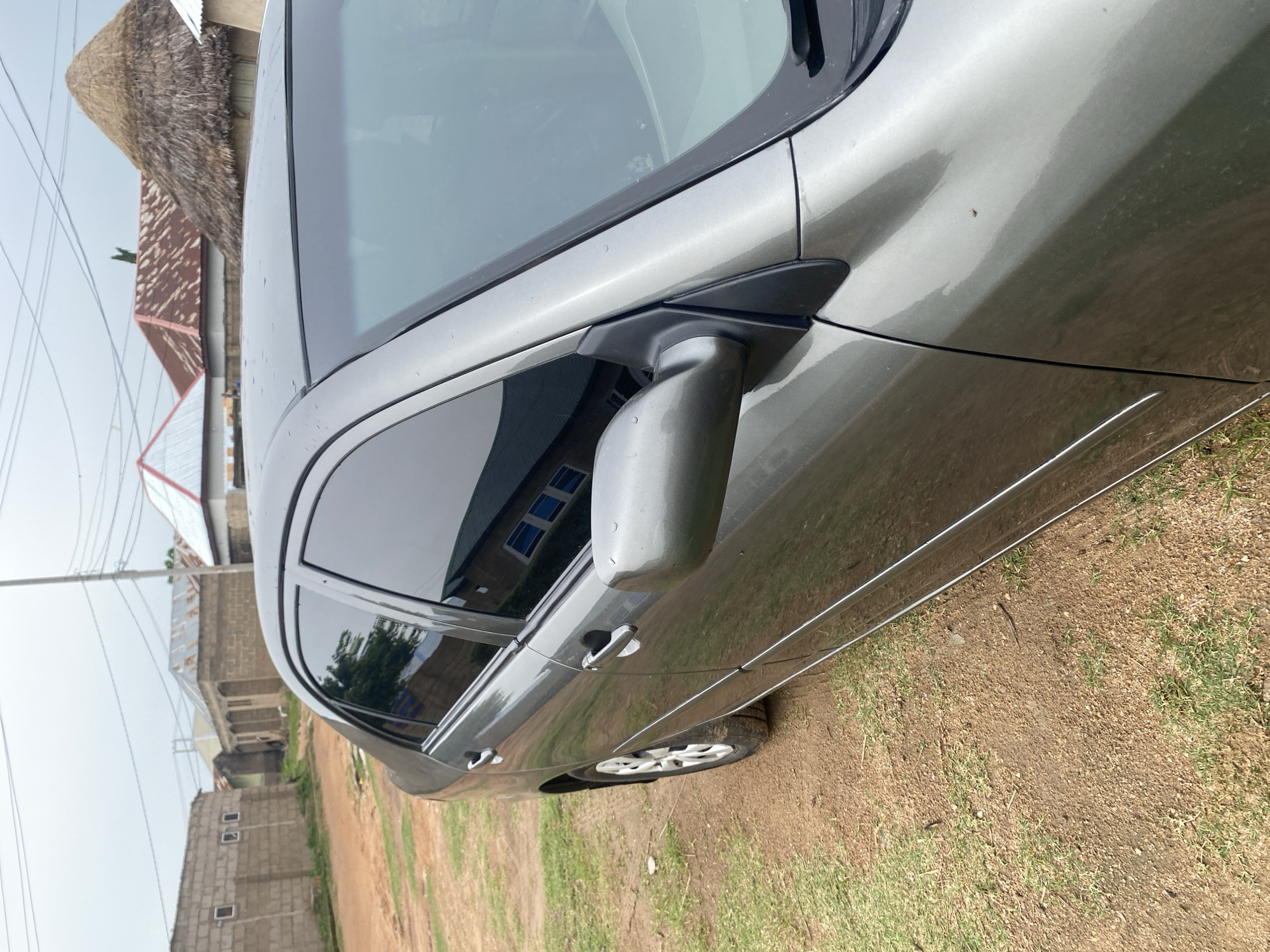 This media file shows a side view of a gray sedan parked on a dirt and grass area. The car's windows are tinted, and the side mirror is visible. In the background, there are buildings with corrugated metal roofs and brick walls, characteristic of a residential area in Jauro Yinu, Nigeria. A power line and utility pole are also present. The sky is overcast, suggesting a cloudy day. The ground is a mix of dry dirt, patches of green grass, and some small stones. There are no people visible in the image. The scene appears to be a typical daytime view in a semi-rural or developing urban setting.