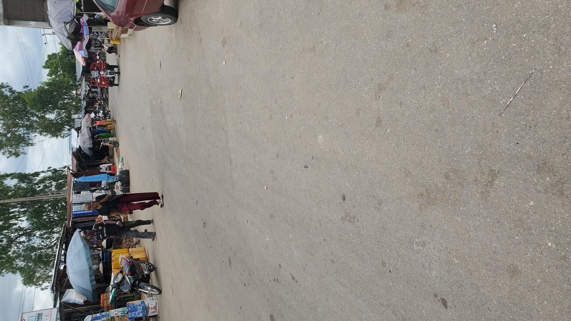 The image depicts a street scene in Jalingo, Nigeria, likely a market area. The foreground is dominated by a paved road with scattered debris and stains. On the left, a black motorcycle is parked near a stall with various goods, including what appear to be yellow containers and stacked items under a blue umbrella. Two individuals are walking towards the right of the frame. The person in front is a male wearing a black t-shirt with "SHARP" visible on it, a baseball cap, and dark trousers. He is walking with a slight bend in his posture. Behind him, a female is walking, wearing dark trousers and a dark top, with a dark bag slung over her shoulder. She appears to be holding something to her mouth. Further down the street, more stalls with umbrellas and merchandise are visible, along with other people and vehicles, including a red three-wheeled auto-rickshaw. The background shows trees and a cloudy sky. The overall atmosphere suggests a busy commercial environment.