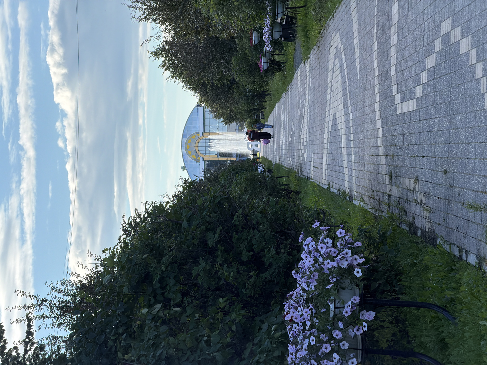 The image shows a paved walkway lined with trees and greenery. In the distance, a large, arched glass building with a yellow accent is visible, with a fountain spraying water in front of it. Two people are walking on the path towards the building. The sky is partly cloudy, suggesting it is daytime. Several planters with purple flowers are placed along the walkway. The overall scene appears to be a park or outdoor public space, possibly an entrance to a botanical garden or attraction. There is no visible text in the image.