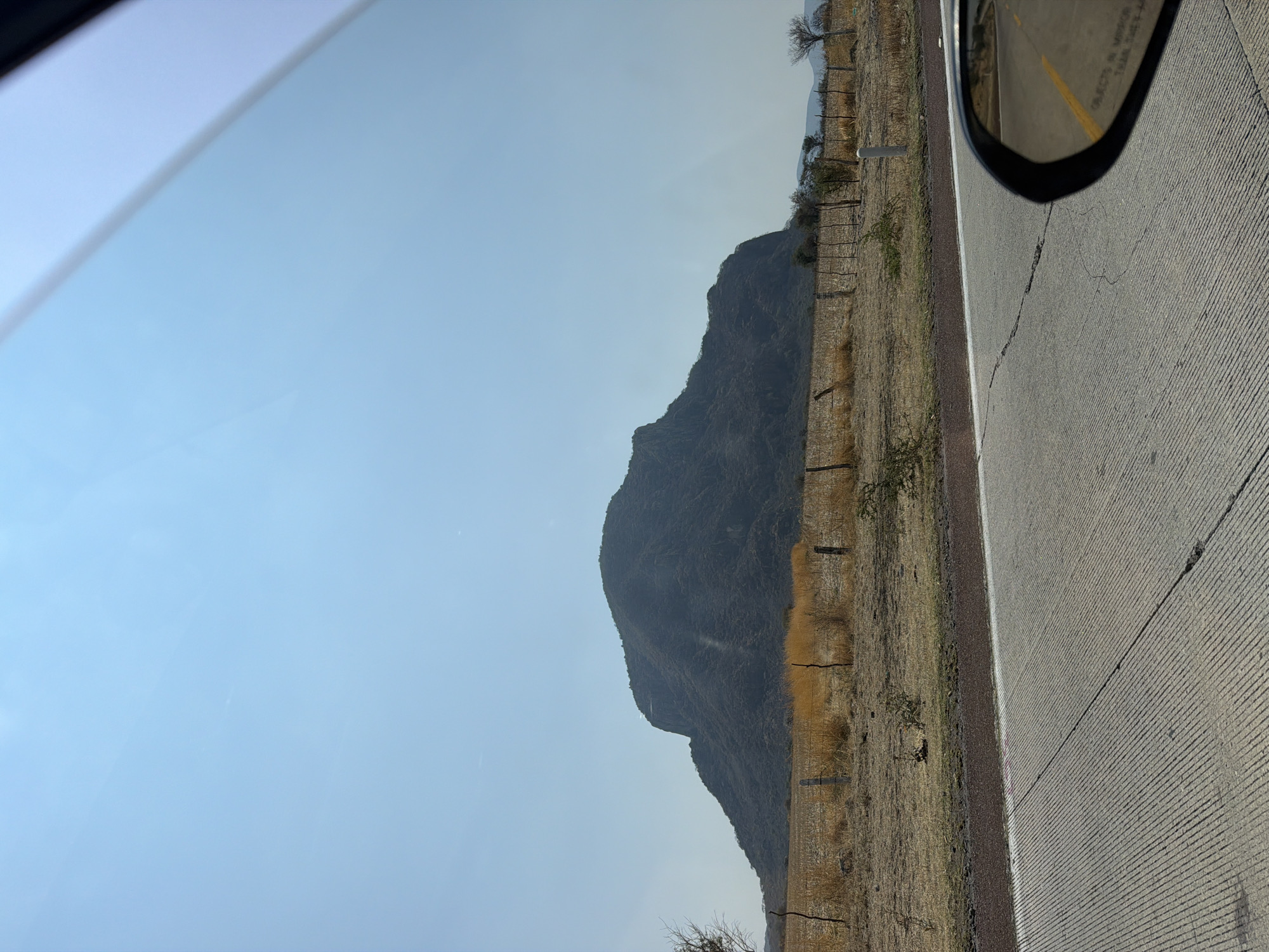 The image depicts a landscape in Jantetelco, Mexico, observed from inside a vehicle. The scene is split between the sky on the left, and the ground on the right. A mountain is located between the two, and a road and a car mirror are seen on the right of the image. The road appears to be made of concrete and has a yellow line separating lanes. The ground near the road consists of dry grass and vegetation, separated by a wooden fence. The sky is clear and blue.