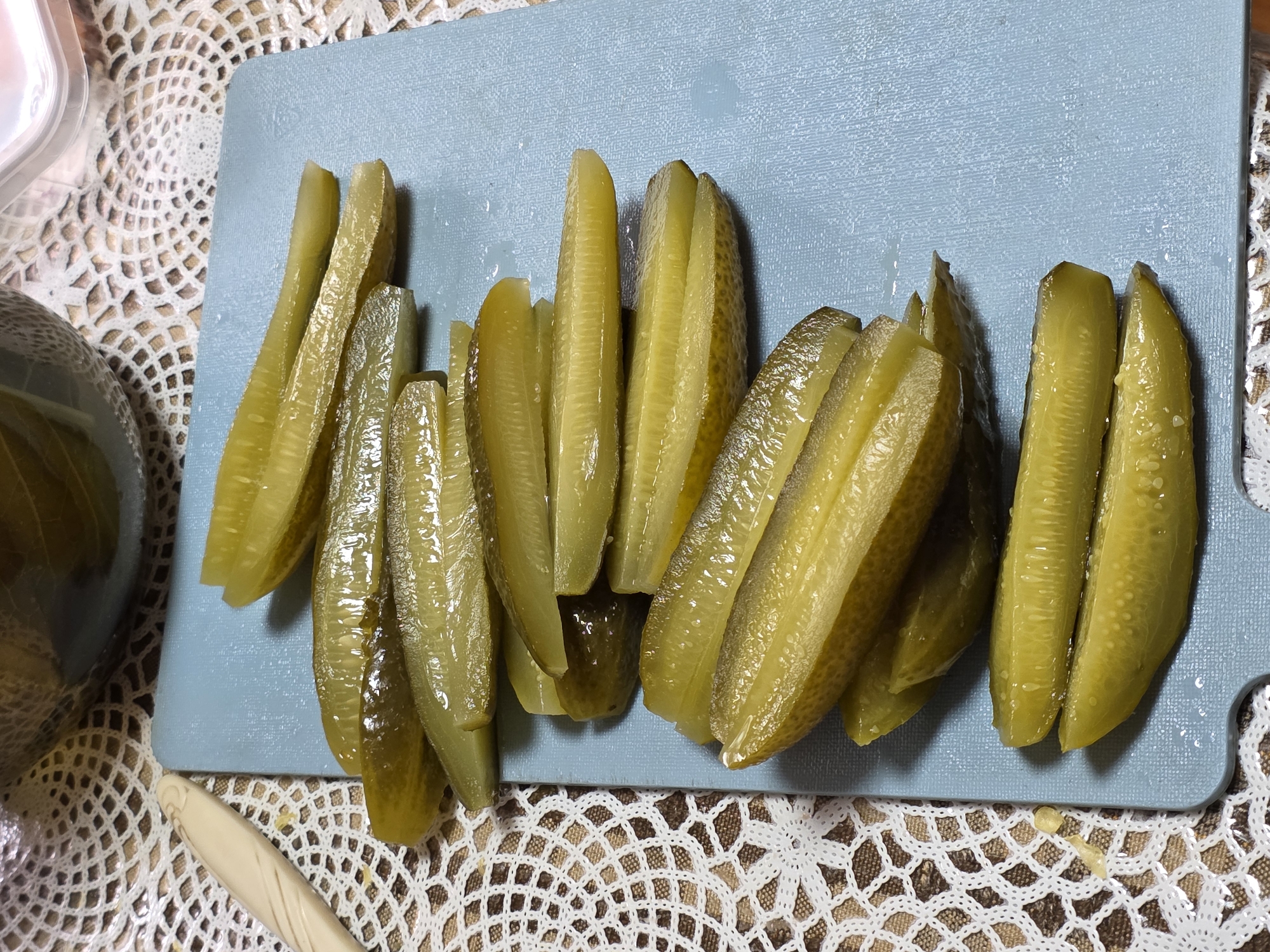 This close-up image, captured in Chaikovsky, Russia, displays numerous sliced dill pickles arranged on a light blue cutting board. The pickles, cut lengthwise into spears or quarters, exhibit a glossy, moist texture and range in color from yellowish-green to olive green. Their bumpy skin is visible on many pieces.
The cutting board itself has a subtle textured surface and some residual moisture from the pickles. It is placed on a white, intricate crocheted or lace tablecloth, which forms a decorative background for the food preparation. In the bottom-left corner, a partial view of a dark glass jar or container is visible, likely the source of the pickles. A small section of a light-colored, possibly wooden or plastic, utensil is also present at the bottom center.
The scene suggests a domestic setting, focusing on the act of preparing or serving food. No individuals are visible, nor is any text present. The lighting is even, indicating an indoor environment.