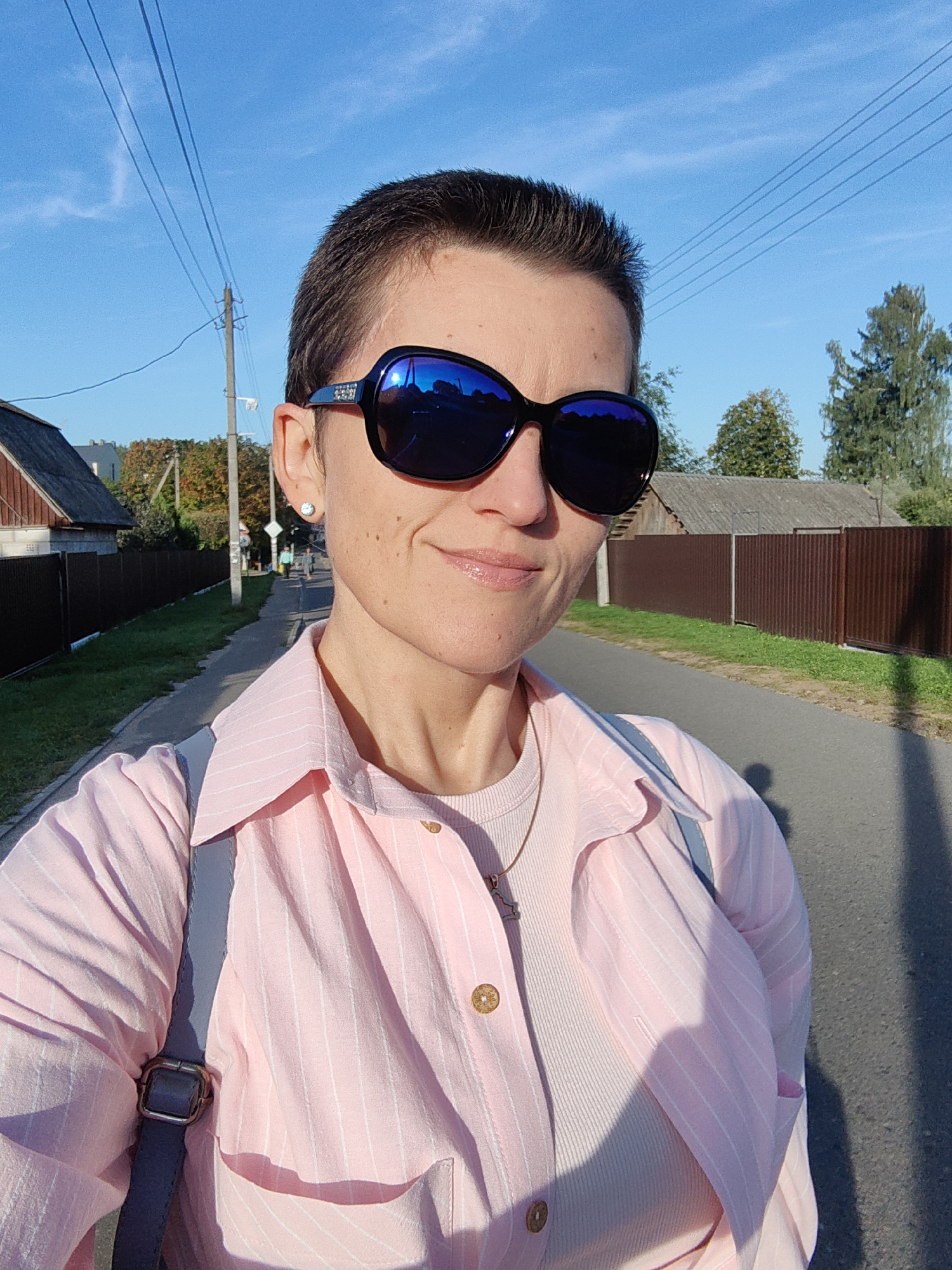 A woman with short dark hair and sunglasses is captured in a selfie outdoors on a paved road. She is wearing a pink striped collared shirt over a light-colored ribbed top and a necklace. A grey strap from a bag is visible across her shoulder. In the background, a road stretches into the distance, flanked by a grassy area and a brown corrugated metal fence. Two houses with dark roofs are visible on the left side of the road. Trees and utility poles with power lines are also present. The sky is clear and blue, indicating a sunny day. The location is identified as Fanipal, Belarus.