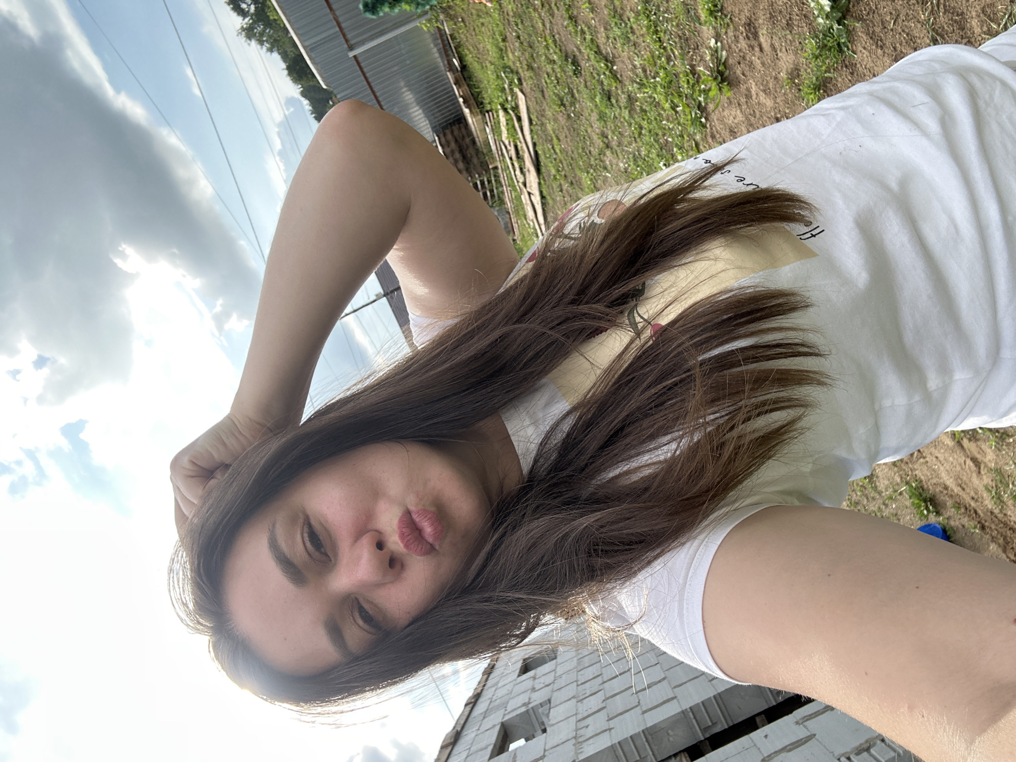In the city of Chaikovsky, Russia, a young woman with long brown hair is taking a selfie outdoors. She is wearing a white t-shirt with a subtle graphic design. Her right arm is raised, with her hand resting on her head, and she is puckering her lips for the camera. The background shows a partly constructed building made of white bricks on the left, and a metal fence and some stacked wooden planks on the right. The ground is covered with dry grass and dirt. The sky above is partially cloudy with the sun shining through.