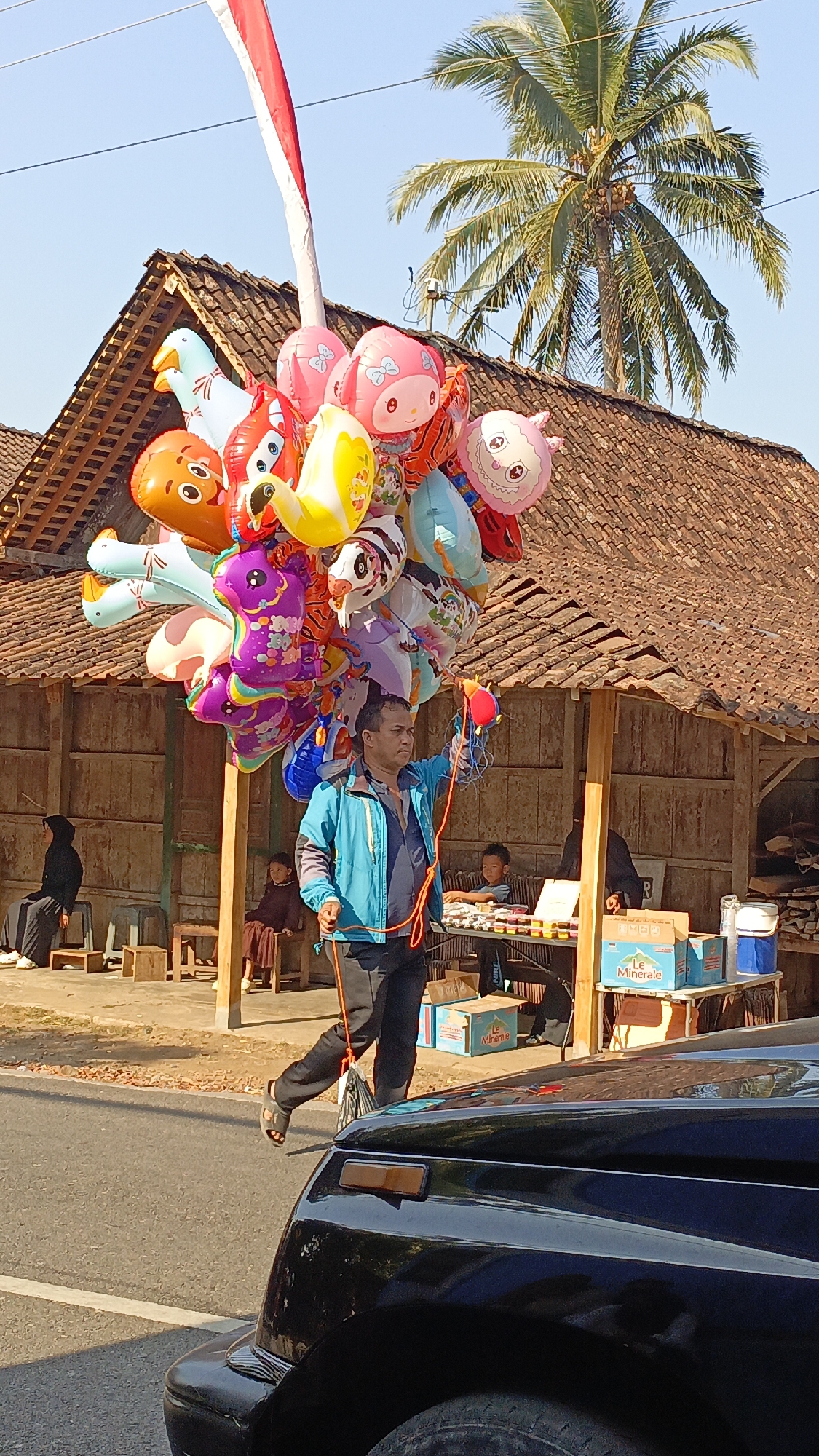 A man walks along a street in Krajan Lor, Indonesia, carrying a large cluster of colorful balloons. The balloons are shaped like various cartoon characters and animals, including a pink Hello Kitty, a yellow duck, a fox, and a purple creature with flowers. The man is wearing a blue jacket and black pants and is holding a red and orange string attached to the balloons. He is walking past a roadside stall with a thatched roof, where several people are seated. A black car is partially visible in the foreground. The background features a coconut tree and traditional Indonesian houses. The scene is brightly lit by sunlight.