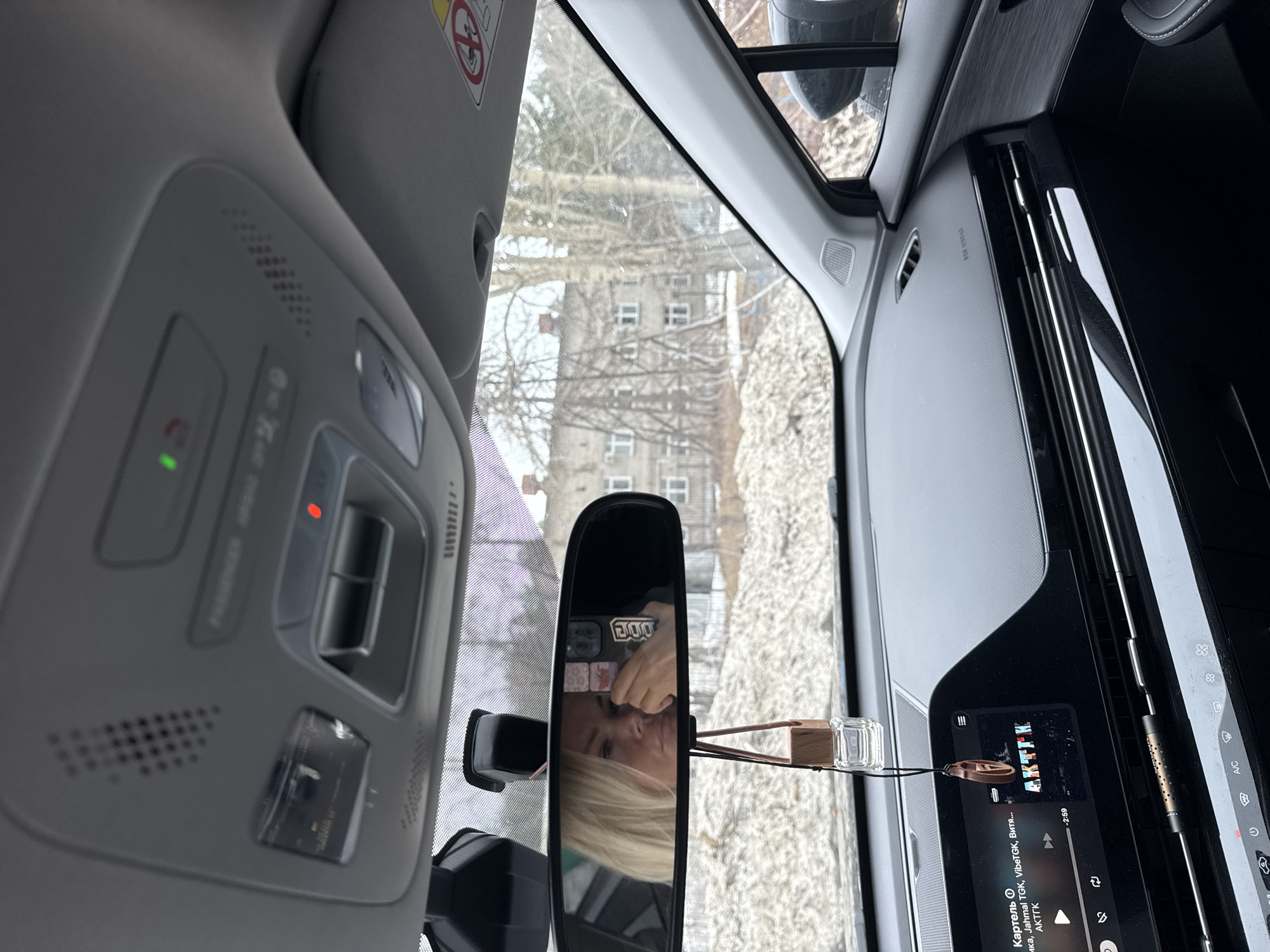 Here is a detailed description of the image:
**Setting:** The image is taken from inside a car, looking towards the front.
**Environment:** Outside, it appears to be winter, with trees visible through the windshield and in the rearview mirror. There is a multi-story building, possibly an apartment building, also visible through the windshield. The location in the city of Unknown, Unknown is determined by the fact that the current song playing on the car's media system is titled "Картель", with the artist listed as "AKTΓK" which suggests a Russian or Slavic origin for the location of the photograph.
**Objects:**
* Car Interior: The interior of the car is visible, including the roof controls, sun visor, dashboard, and center console screen.
* Rearview Mirror: The rearview mirror reflects the photographer. There is an air freshener hanging near the rearview mirror.
* Media Screen: The media screen displays the song information and controls.
* Smartphone: A smartphone can be seen through the rearview mirror.
**People:**
* A woman is visible in the rearview mirror. She appears to be holding a phone, possibly taking the picture.