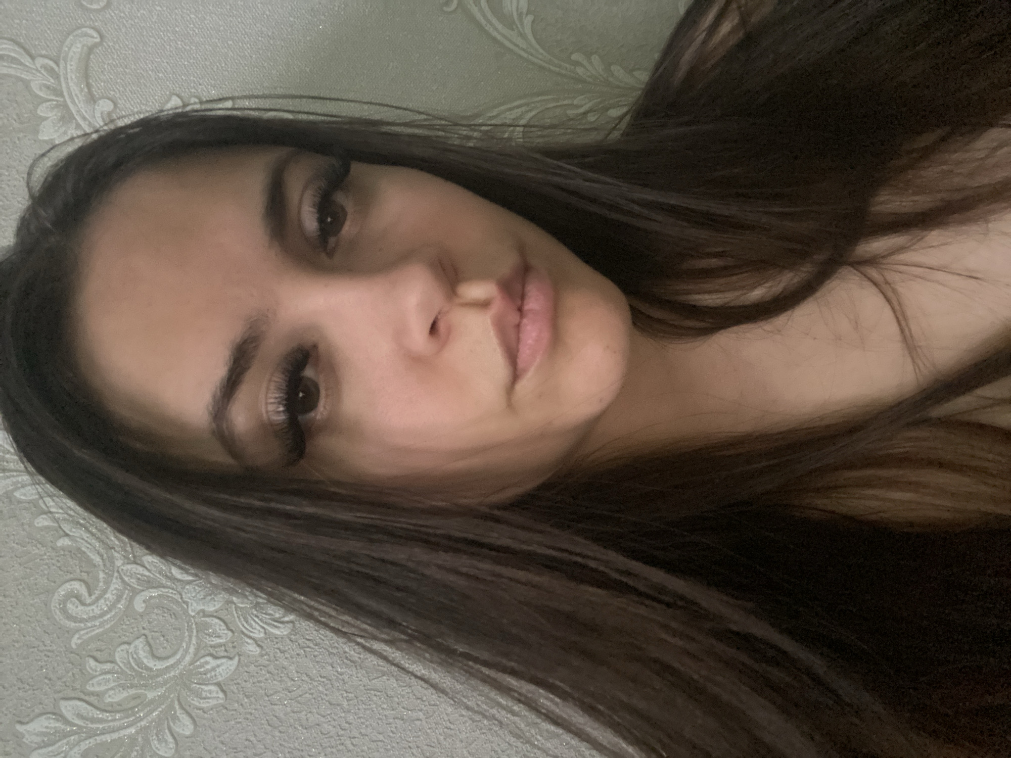 A selfie of a woman with long, dark brown hair and brown eyes, with prominent eyelashes, is taken at a slight angle. Her skin tone appears fair. The background shows a light-colored wall with a floral pattern. The image's location is unknown and cannot be determined from the provided information, which does not include specific details about Chaikovsky, Russia.