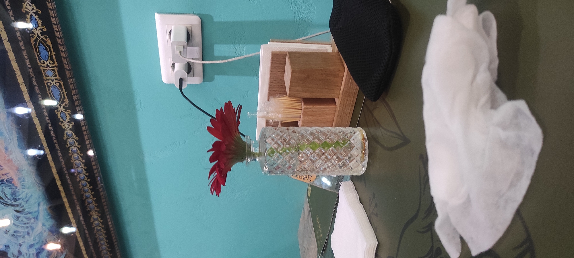 A close-up indoor shot captures a table setting with a vibrant red gerbera daisy in a patterned glass vase as the focal point. The vase sits on a dark green surface adorned with a subtle leaf pattern. To the left, a stack of white paper napkins is visible, and further back, a menu is partially obscured. Behind the flower, a wooden organizer holds toothpicks and what appear to be napkins. A black mesh object rests on the table to the right of the organizer. In the background, a turquoise wall features a white electrical outlet with two white adapters and charging cords plugged in. The top of the image reveals a decorative border with intricate gold and blue detailing, above a framed artwork. The lighting suggests an indoor environment, possibly a cafe or restaurant. No people or animals are present in the scene. The image does not contain enough visual information to determine its location in Moscow, Russia.