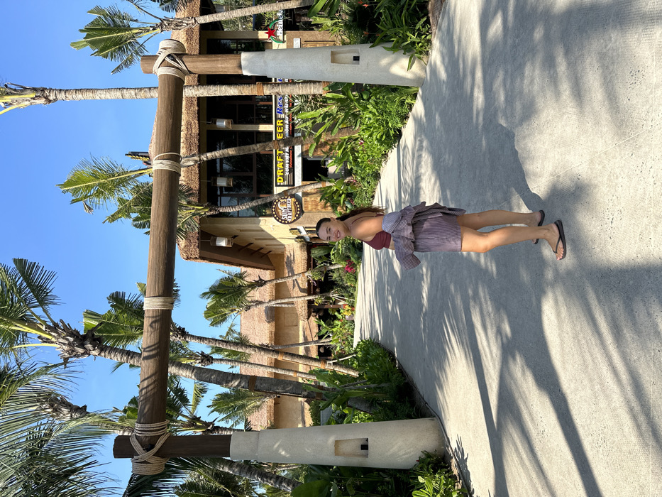 A female subject stands on a light-colored paved pathway under a clear blue sky, looking over her right shoulder towards the camera, at the entrance to a resort or commercial complex in Sunset Town, An Thới Ward, Phú Quốc City, Kiên Giang Province, Vietnam. She has dark hair tied back and is wearing a maroon spaghetti strap top, dark gray striped shorts, and a light-colored striped shirt draped over her shoulders, paired with dark flip-flops.
In the immediate foreground, a rustic wooden archway, constructed from thick posts and a horizontal beam bound with natural rope, spans the path. The pathway itself curves slightly to the left and exhibits distinct shadows from overhead foliage, indicating bright sunlight. Lush tropical vegetation, comprising various types of green plants and numerous tall palm trees, lines both sides of the path and extends into the background.
Behind the subject, several multi-story buildings with distinctive thatched roofs and wooden facades are visible amidst the palm trees. The most prominent building displays large windows and multiple illuminated signs, including a circular yellow sign for "SUM'S KRAFTEERS" and a rectangular sign reading "D'RAFT BEER Restaurant VÒM VIỆT PHÚ QUỐC." A green Heineken logo is also visible on the right side of the building. The overall environment suggests a well-maintained tropical leisure or dining establishment.