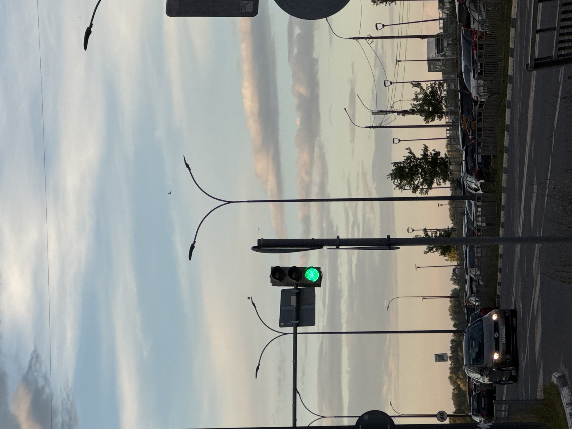 The image depicts a traffic scene at dusk in Nizhny Novgorod, Russia. The photograph is oriented vertically. In the foreground, a traffic light displays a green signal. To the right of the traffic light, a dark-colored car is visible on the road, its headlights illuminated. Further down the road, more vehicles are parked along the side. The background shows a line of trees and a sky with wispy clouds, indicating a late evening or early morning time of day with potentially overcast weather. Several streetlights are present throughout the scene, with their black poles and curved arms. The overall impression is of a quiet street or parking area as daylight fades. There is a speed limit sign visible with "40" printed on it.