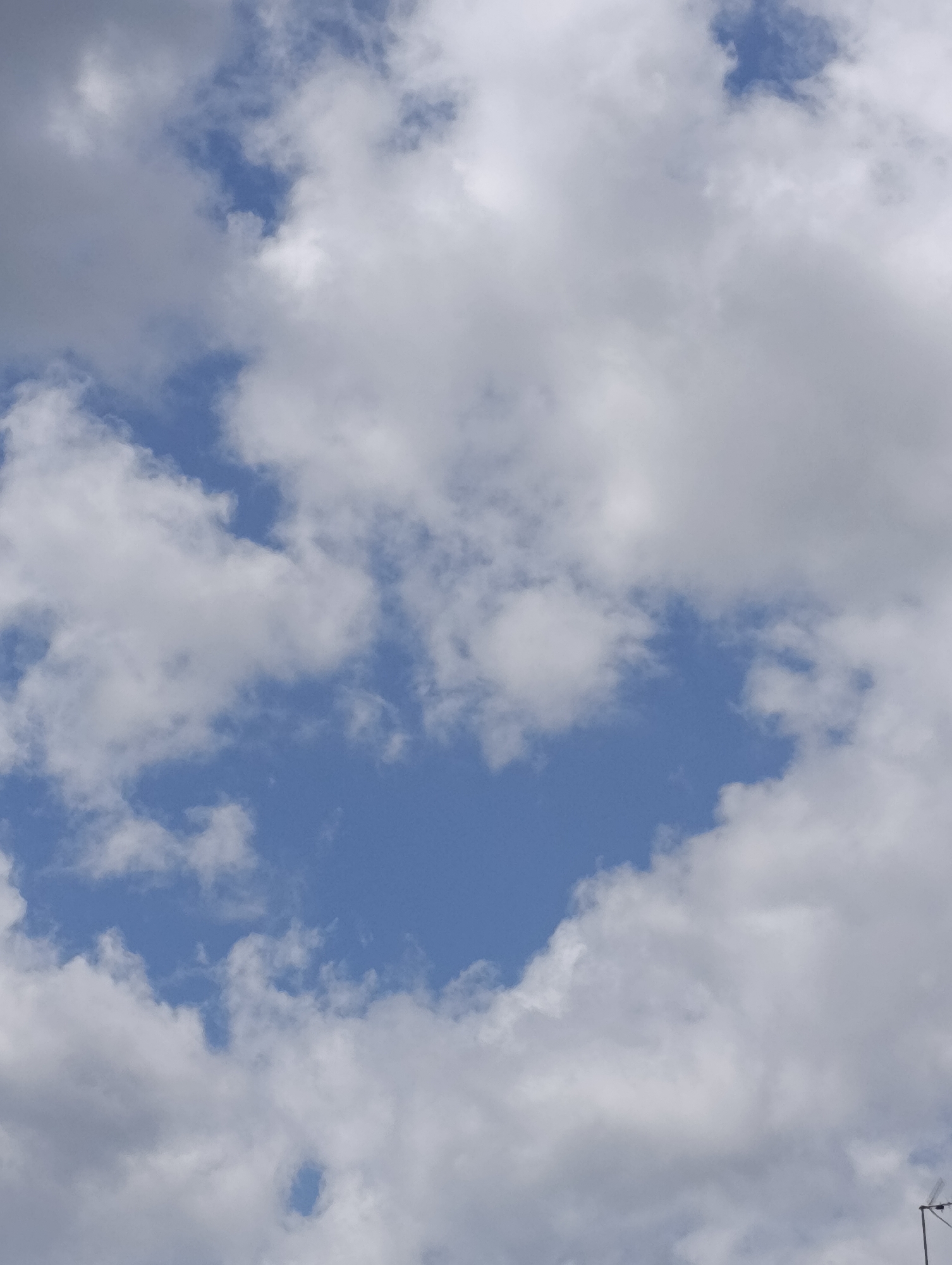A vertical, close-up, medium-shot photograph captures a partial view of a bright blue sky peppered with fluffy white clouds. The clouds are scattered across the frame, with some appearing dense and opaque, while others are thinner and more translucent, allowing the blue sky to peek through in several areas. The light appears to be coming from above, illuminating the clouds and creating subtle shadows and highlights. In the bottom right corner, a small portion of a street lamp post is visible, with its dark metal structure and a small, angled light fixture extending to the right. The image is devoid of any discernible human presence or activity. The scene is characteristic of an outdoor environment on a partly cloudy day. The specific location in the city of Setúbal, Portugal, cannot be determined from the provided image alone, as it focuses solely on the sky and a small portion of infrastructure.