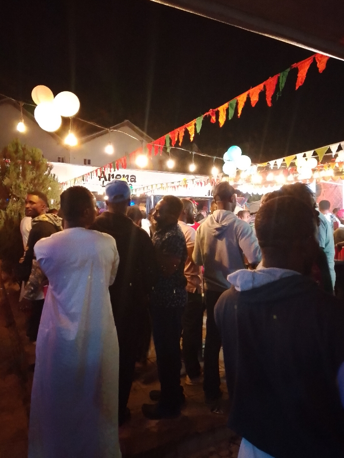 The image depicts an outdoor night scene in Narayi, Nigeria, featuring a crowd of individuals. Predominantly male, attendees are gathered under decorative lighting, which includes multiple strings of warm white spherical and teardrop-shaped bulbs, as well as several large, round white globe lights. Overhead, strands of green, orange, and red triangular pennant flags are visible. A cluster of light blue and white balloons is present in the upper right.
In the foreground, a person in a long, white traditional robe is visible from behind on the left, and another person in a dark hoodie is seen from behind on the right. Individuals in the midground wear casual attire, including a blue baseball cap, a grey hoodie, and a dark patterned shirt, all facing towards a large white tent or canopy marked "Arena." Behind this structure, a white building with a dark roof is discernable. A dark green, bushy plant is situated on the far left of the frame. The ground appears dark and unpaved, consistent with an outdoor event area. Individuals are standing and mingling within the illuminated space.